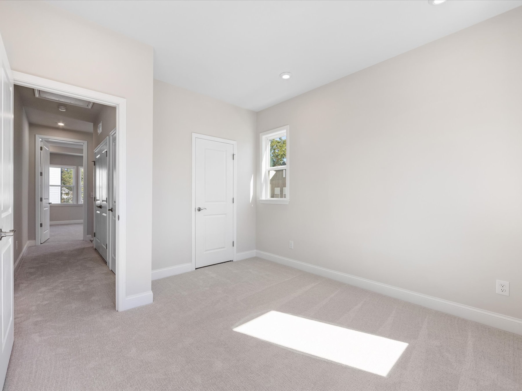 A bright and spacious room with white walls, a hardwood floor, and a doorway leading to another room. The room appears to be a bedroom or living space, with a window allowing natural light to fill the space.