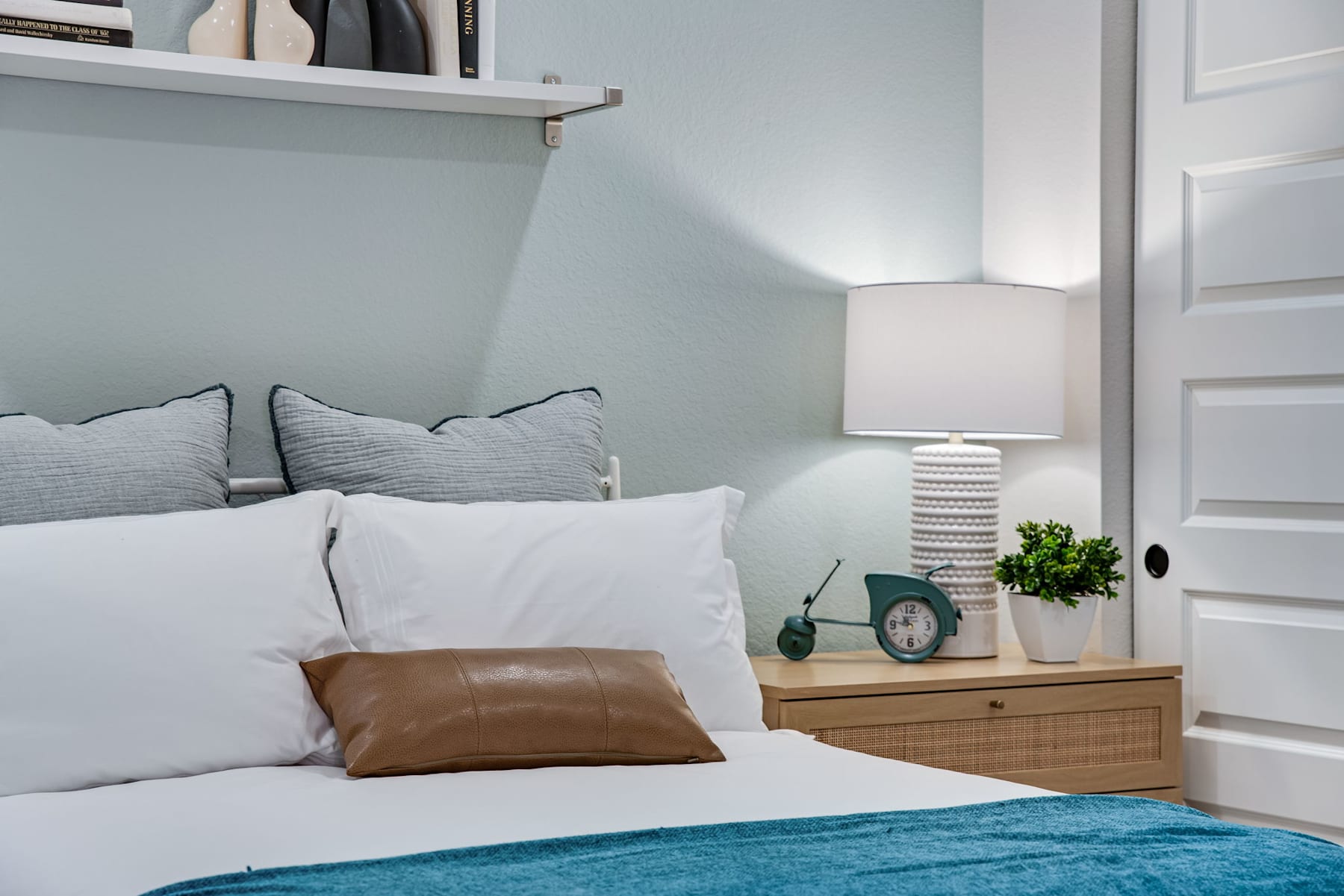 A cozy and inviting bedroom with a white bed, gray pillows, a wooden nightstand, and a potted plant, all set against a light blue wall and a white door.