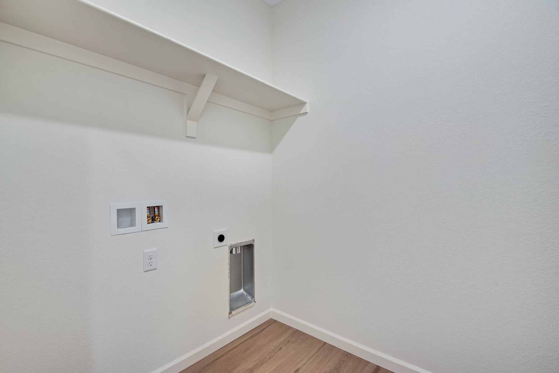 A simple, minimalist room with white walls, a wooden floor, and a small electrical outlet visible in the corner.