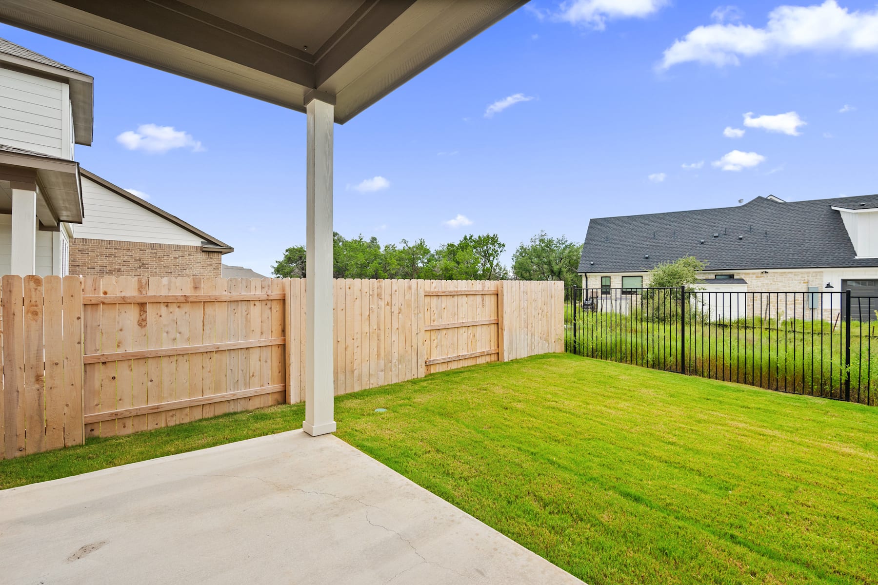 A well-manicured backyard with a wooden fence, a grassy lawn, and a covered patio leading to a residential neighborhood in the background.