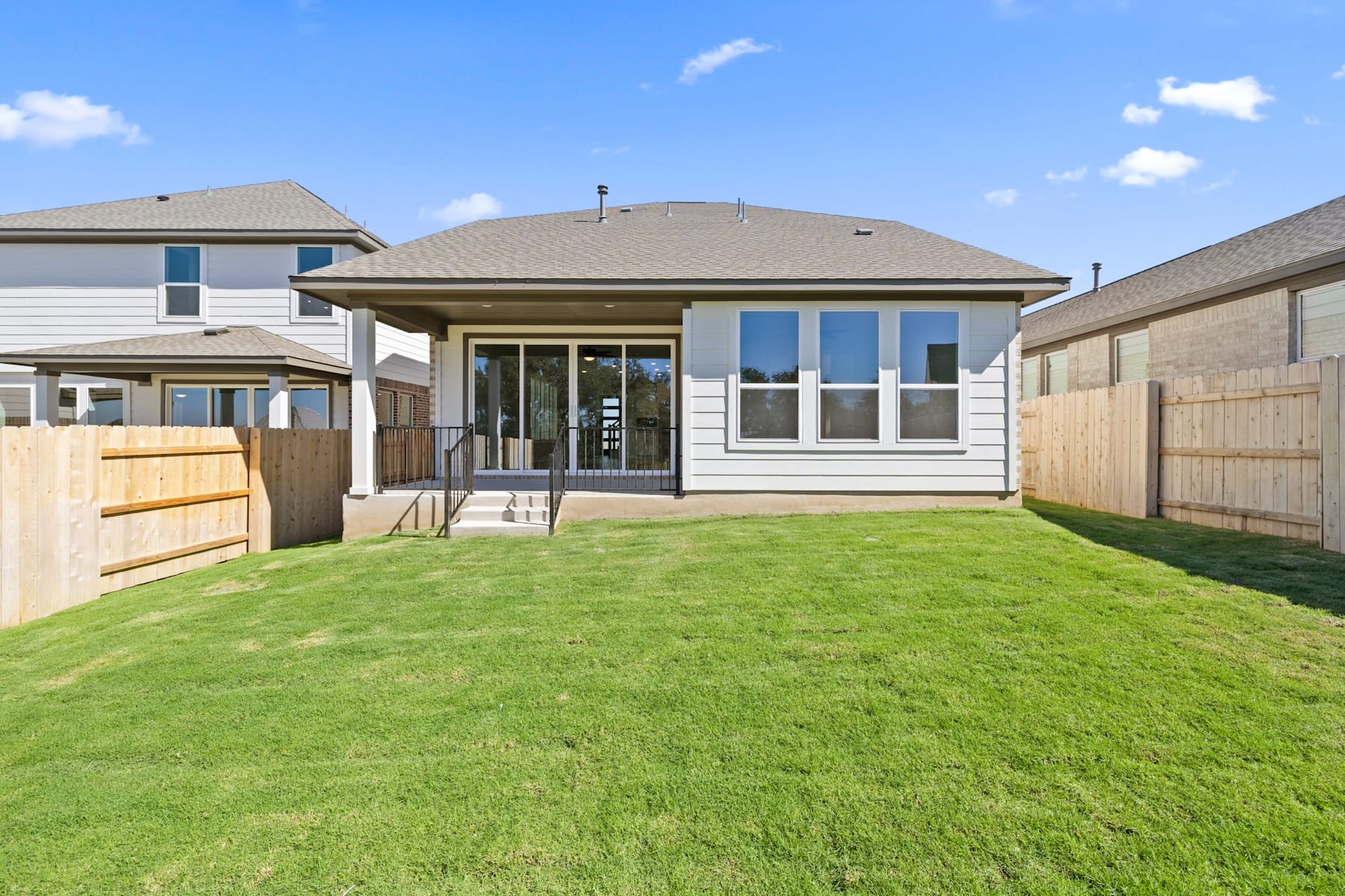 A well-manicured lawn leads to a modern, single-story house with a covered porch and large windows, set against a clear blue sky with scattered clouds.