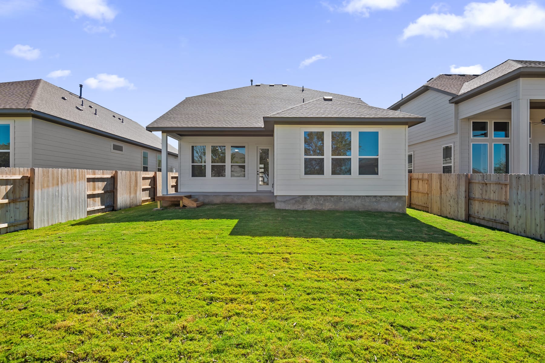 A well-manicured lawn with lush green grass surrounds a modern, two-story residential home with a gray exterior and a tiled roof, set against a clear blue sky with scattered clouds.