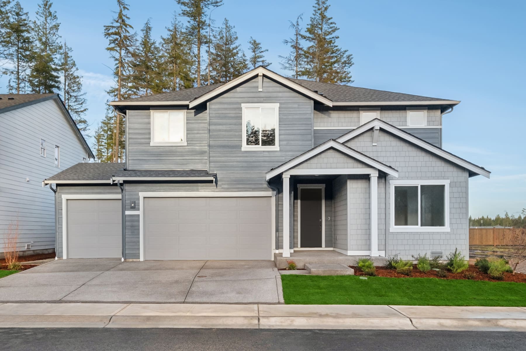A two-story gray house with a garage stands in the foreground, surrounded by pine trees and a grassy lawn in the background.