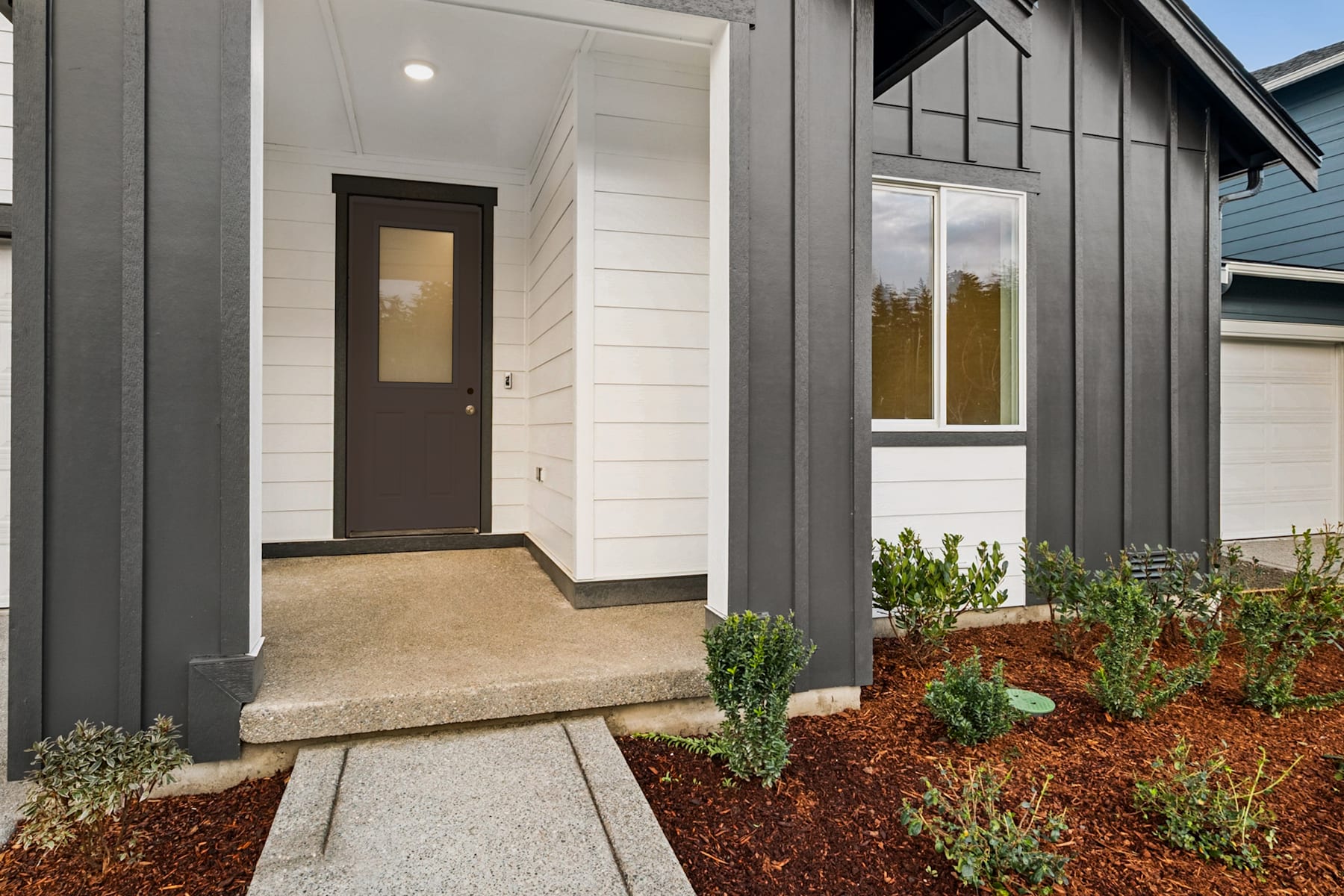 The image shows the exterior of a modern, gray-colored house with a wooden door and a small garden in the foreground, surrounded by a mulch-covered landscape.
