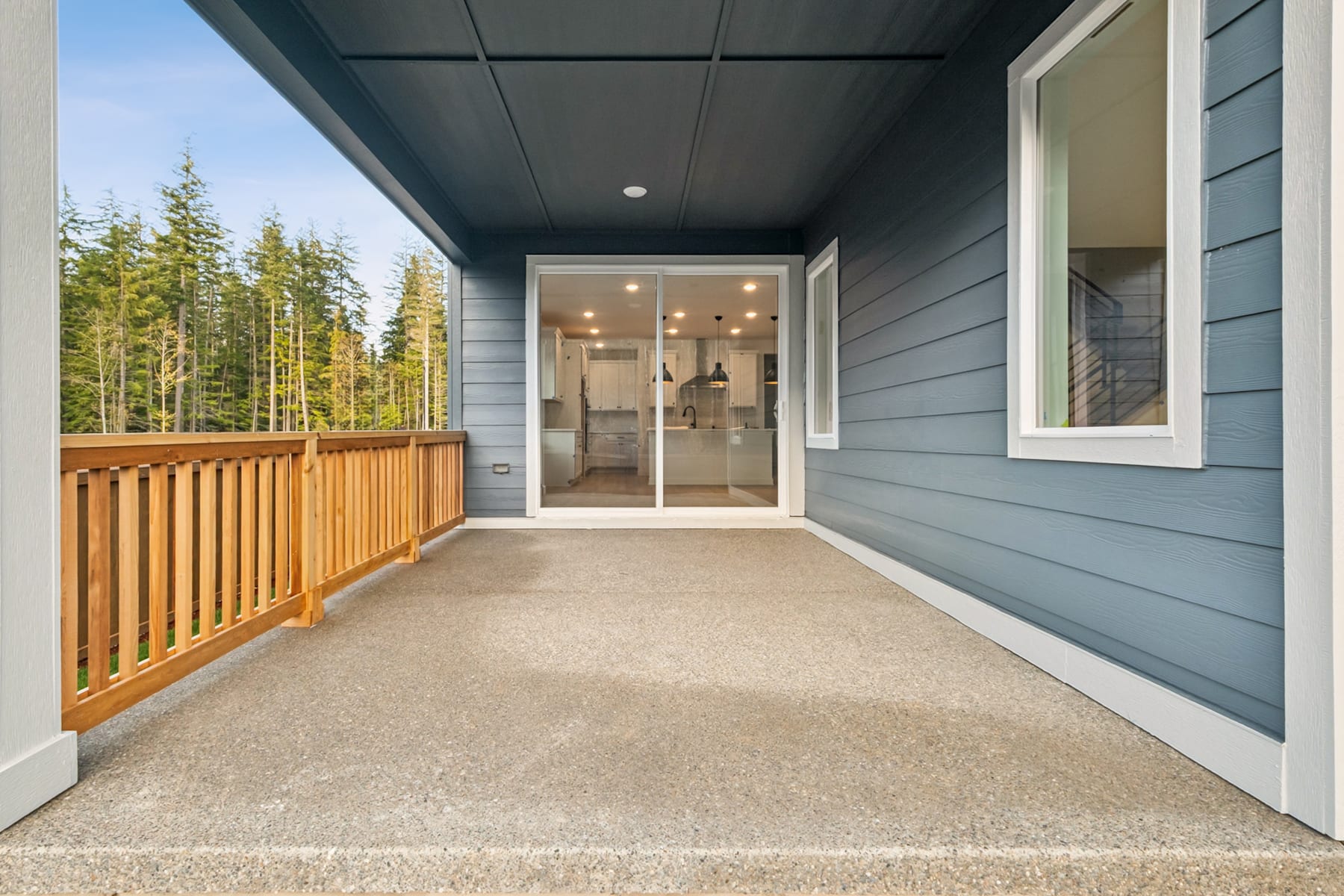 A covered porch with a wooden railing leads to a glass-paneled entryway, surrounded by a forested landscape in the background.