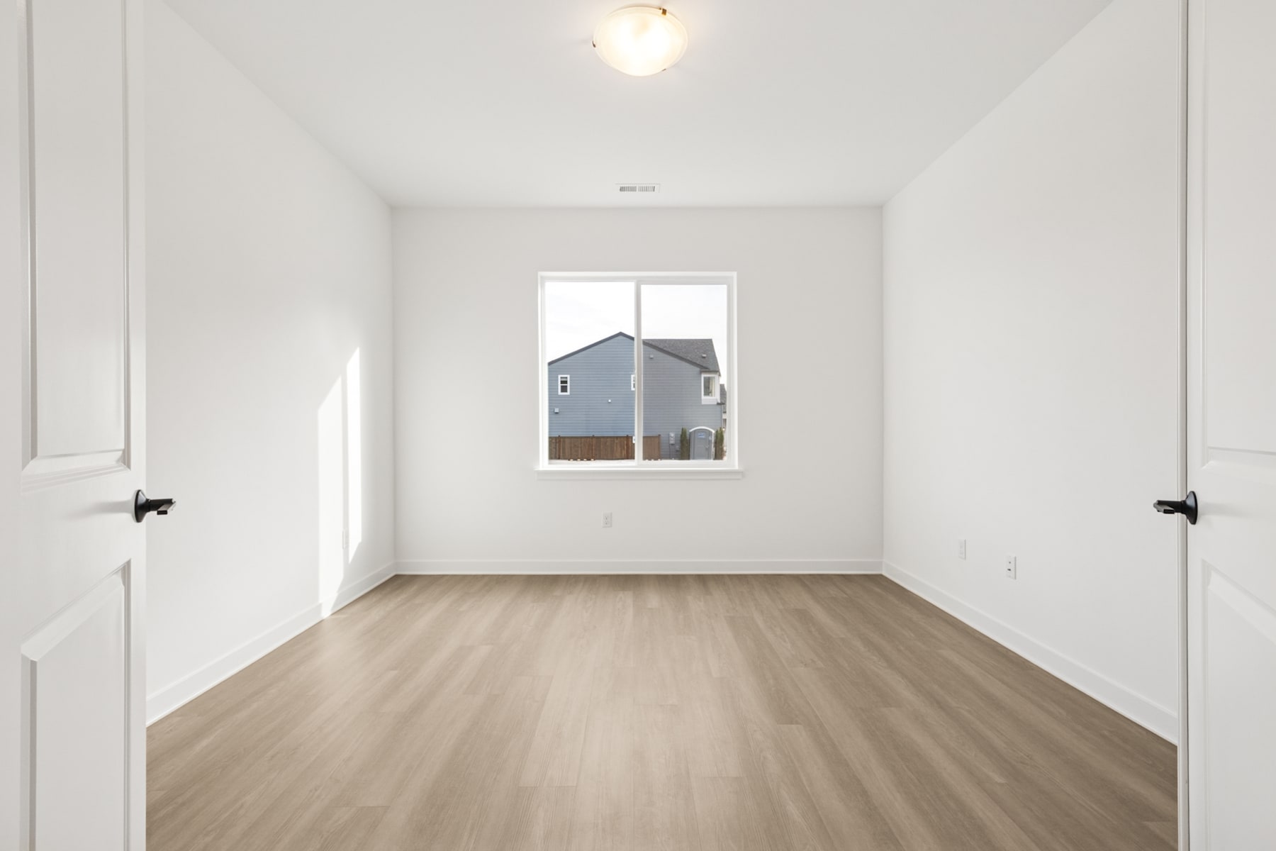 A bright, minimalist room with white walls, a wooden floor, and a window overlooking a residential building in the background.