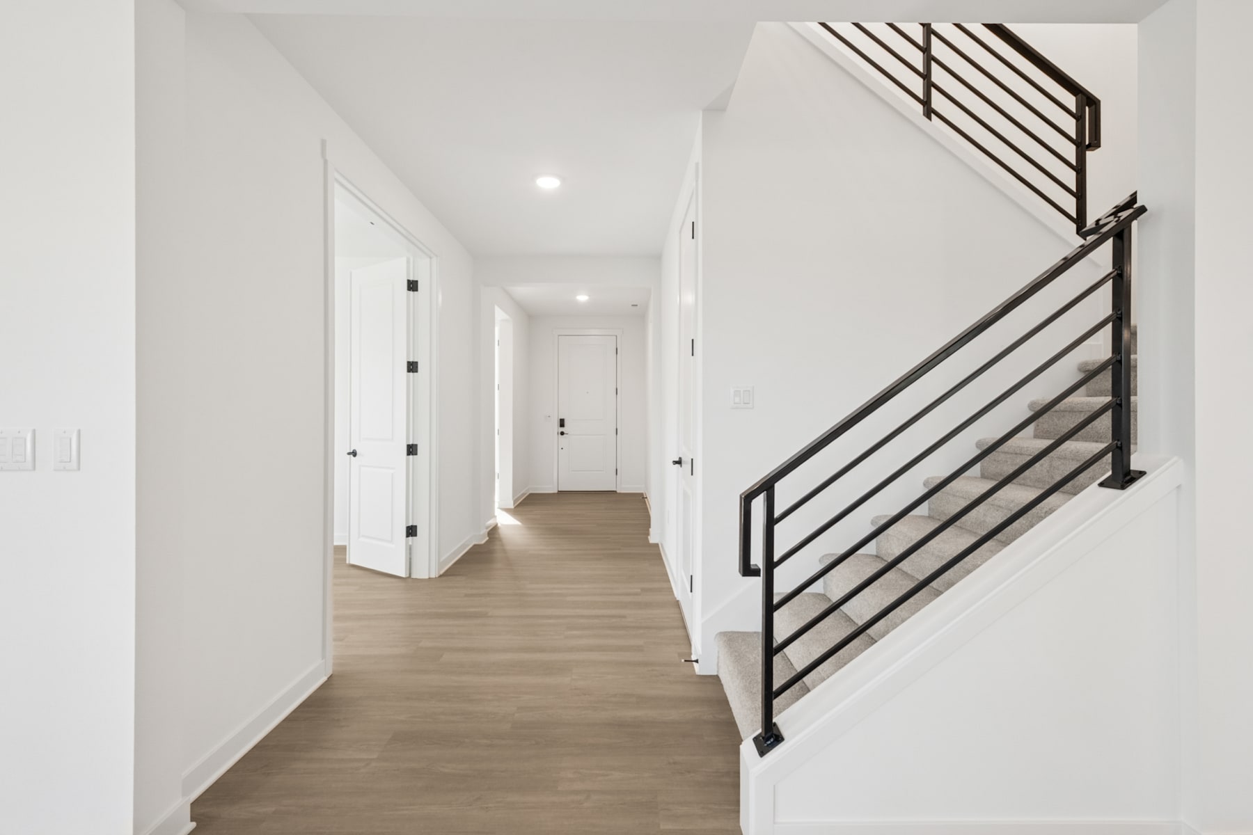 A bright, minimalist hallway with a wooden floor and a modern metal staircase railing leading up to the second floor.