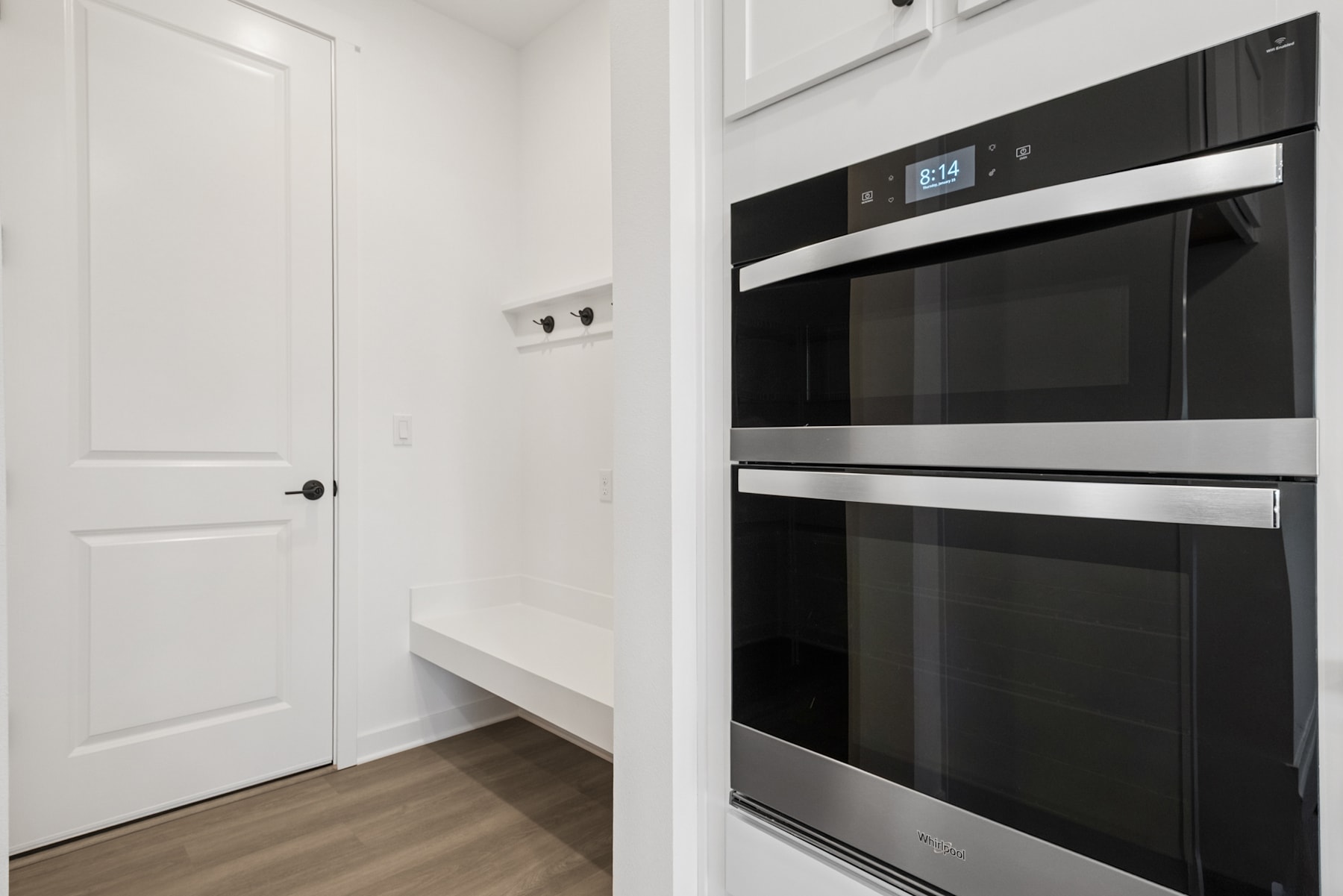 A modern, minimalist kitchen with a sleek black oven and microwave set against a white wall and wooden flooring.