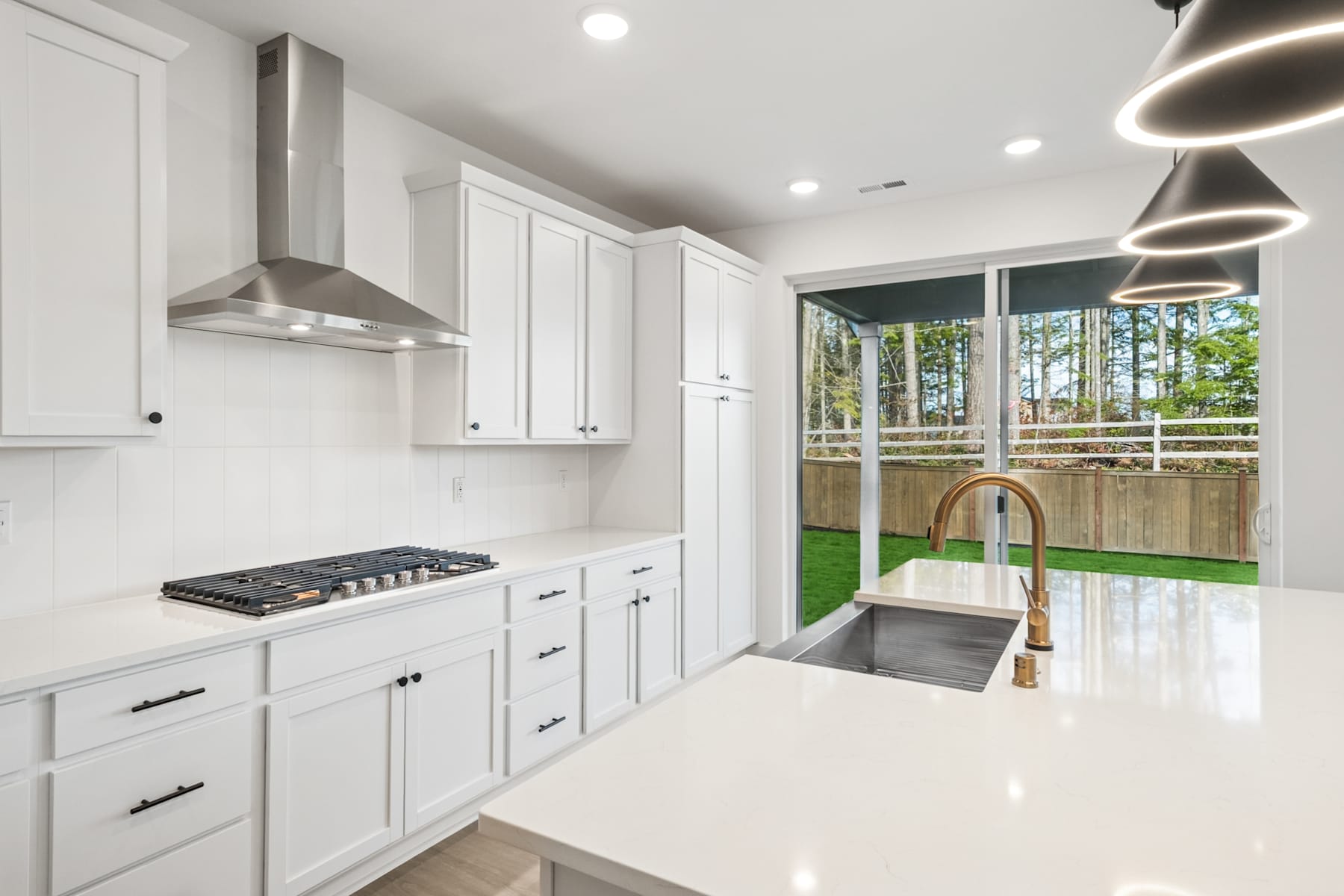 A modern, well-lit kitchen with white cabinets, stainless steel appliances, and a large window overlooking a backyard with trees.