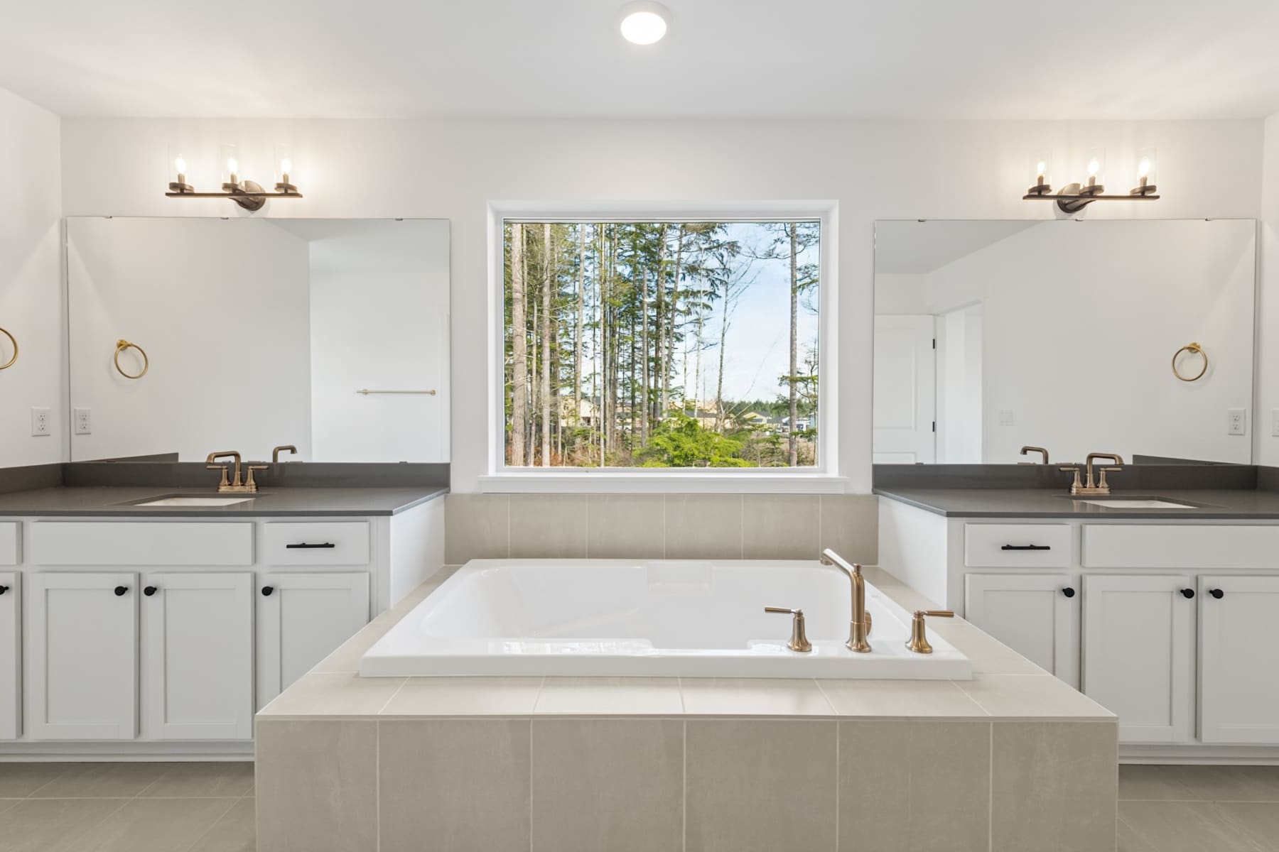 A spacious and well-lit bathroom with a large window overlooking a lush, forested landscape, featuring a freestanding bathtub and dual vanity sinks with modern fixtures.