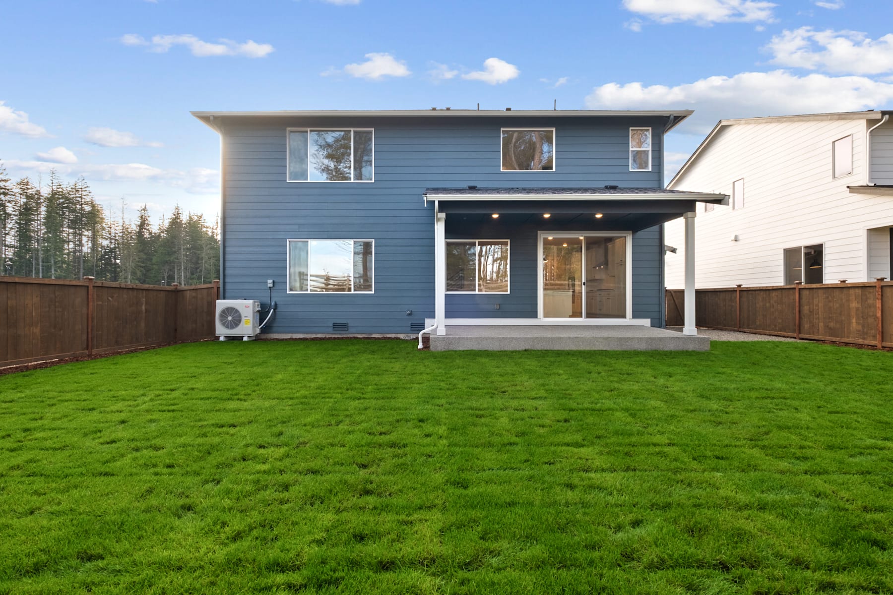A modern two-story house with a blue exterior stands on a lush green lawn, surrounded by trees in the background.