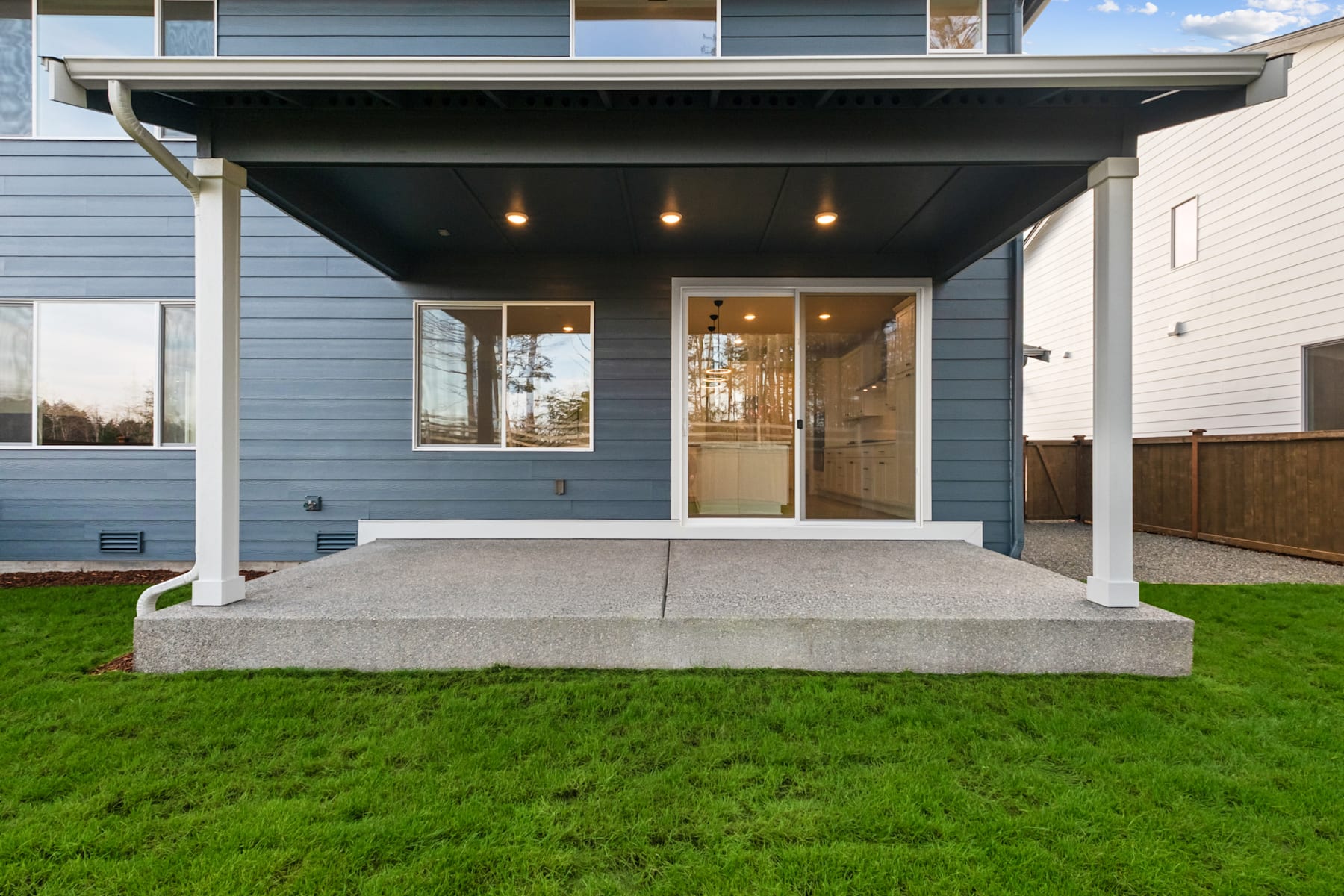 A modern, two-story house with a covered porch, surrounded by a lush green lawn and a wooden fence in the background.