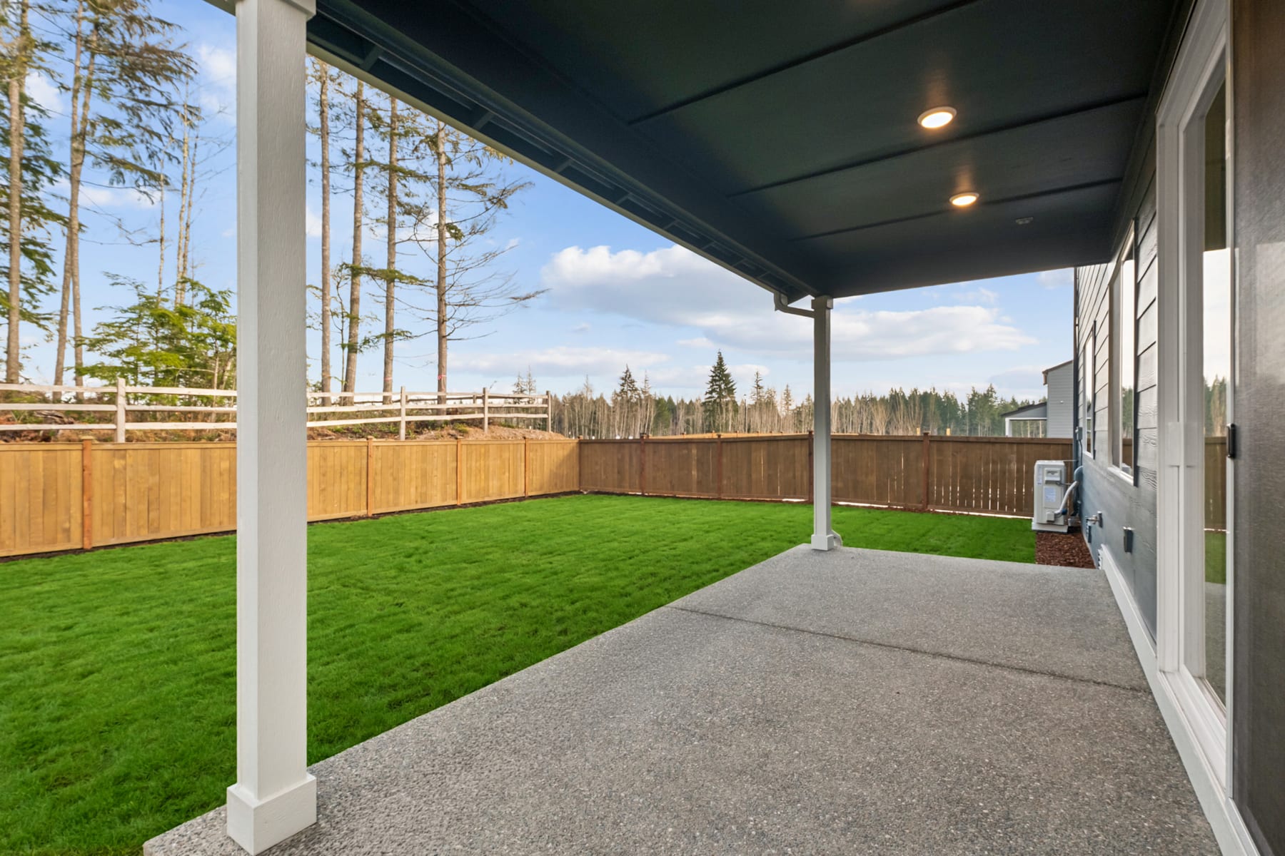 A covered patio with a lush green lawn and a wooden fence in the background, surrounded by tall pine trees.