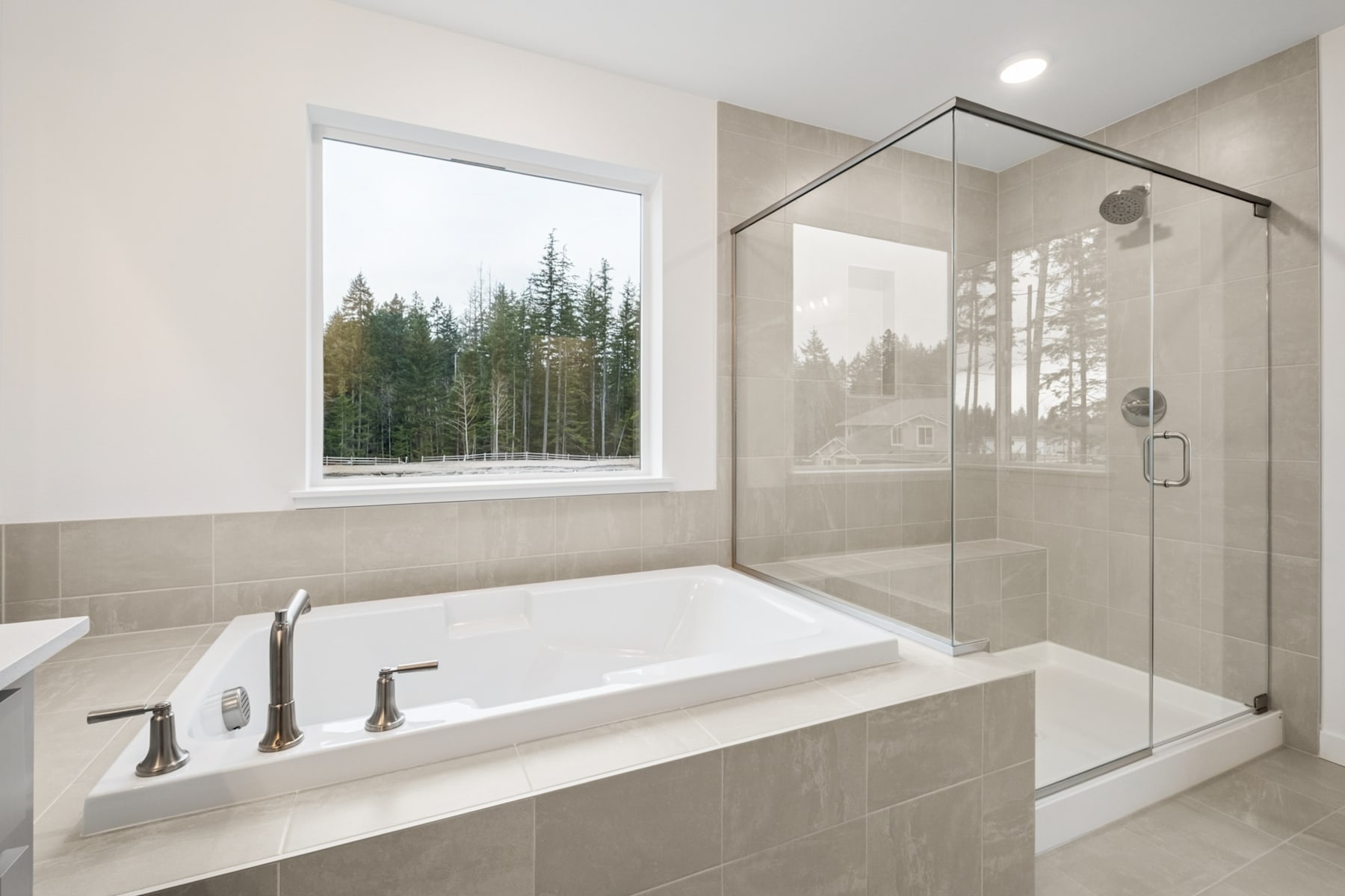 A modern bathroom with a large window overlooking a forested landscape, featuring a bathtub, a glass shower enclosure, and neutral-toned tiles.