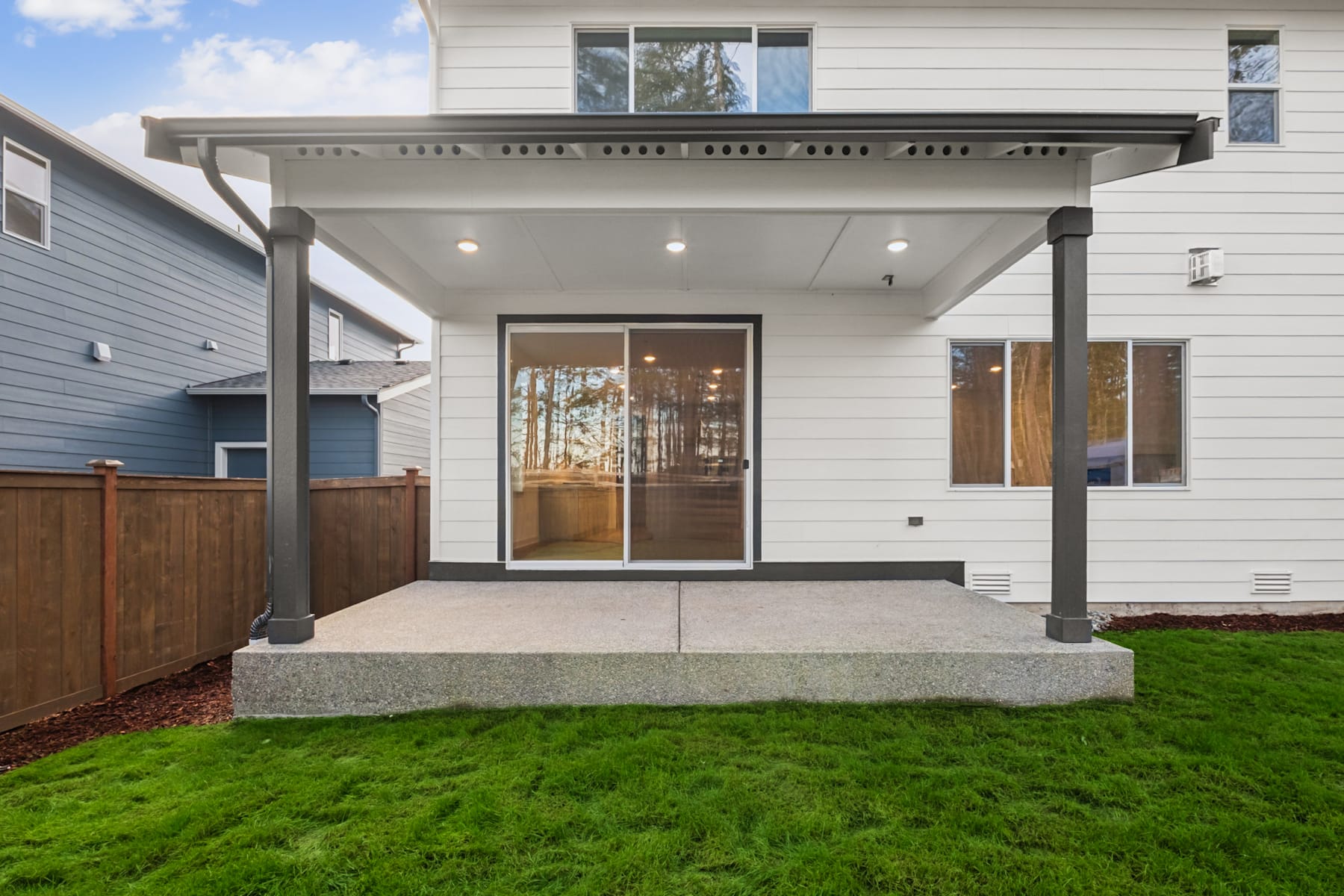 A modern, two-story house with a covered porch, surrounded by a lush green lawn and a wooden fence in the background.