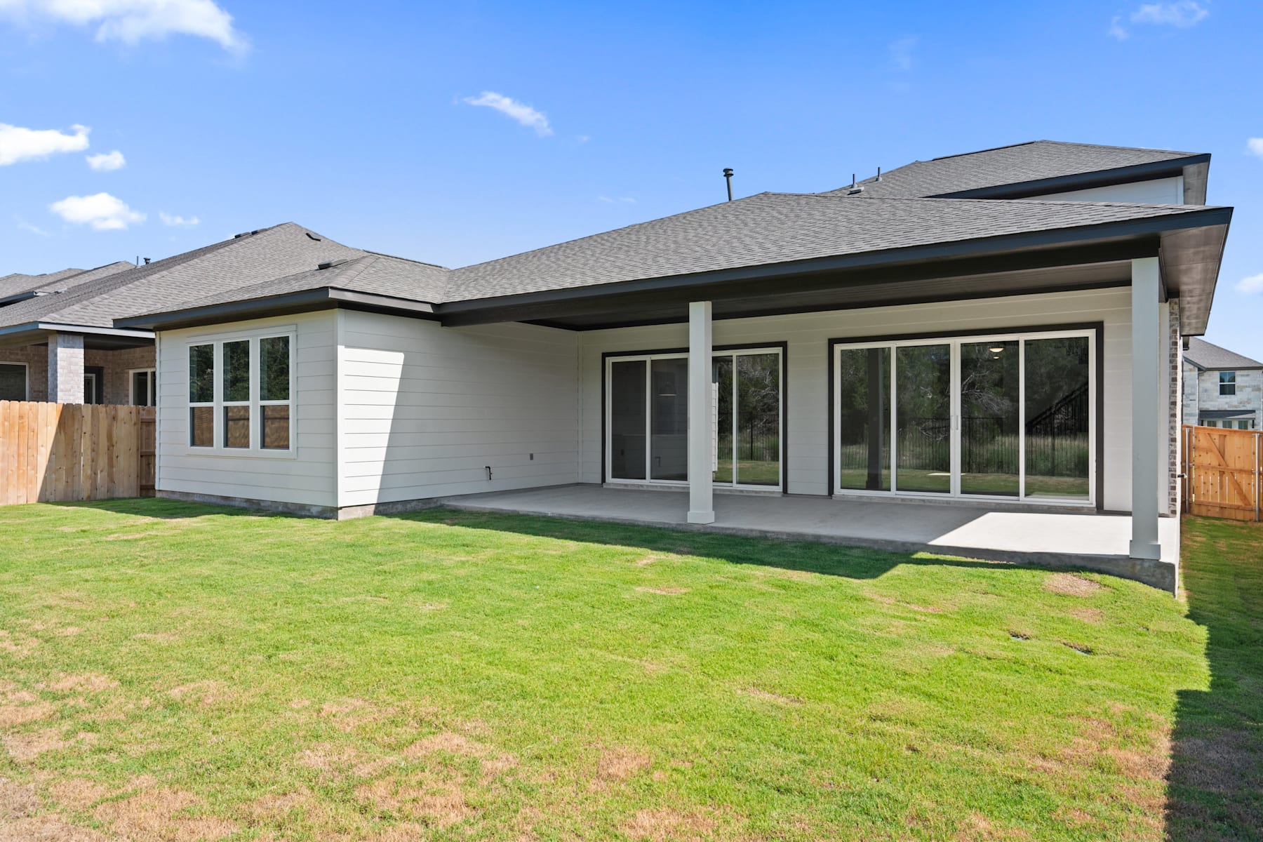 A single-story modern house with a gray roof and white exterior, surrounded by a lush green lawn in the foreground, set against a clear blue sky with scattered clouds.