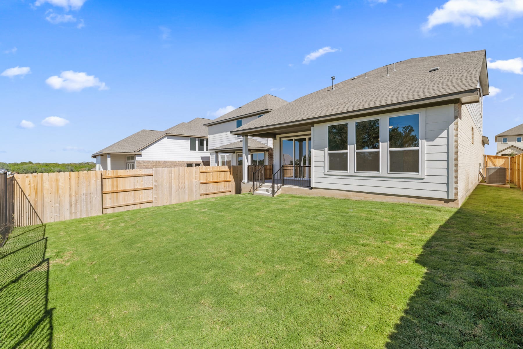 A well-manicured lawn surrounds a modern, two-story residential home with a wooden fence and a clear blue sky with fluffy clouds in the background.