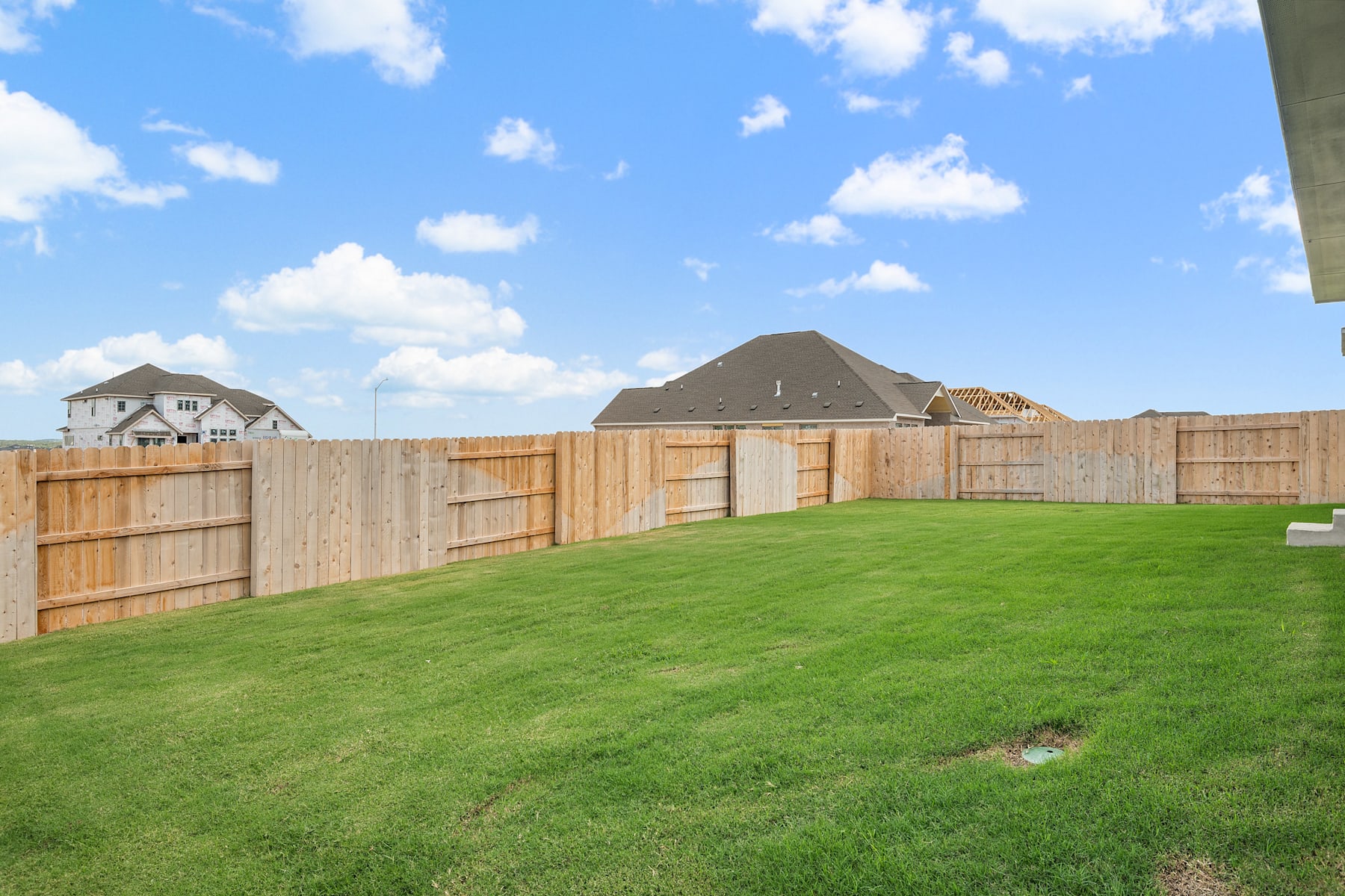 A well-manicured lawn surrounded by a wooden fence, with houses visible in the background against a blue sky with fluffy white clouds.