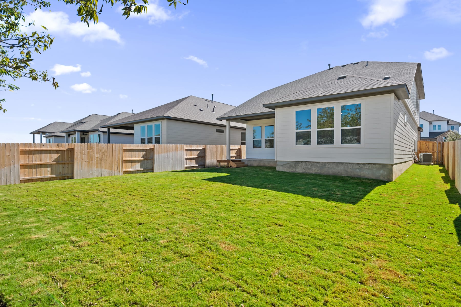 The image shows a residential neighborhood with several modern, two-story houses surrounded by well-manicured lawns and fenced backyards. The foreground features a lush, green lawn, while the background showcases a clear blue sky with scattered clouds.