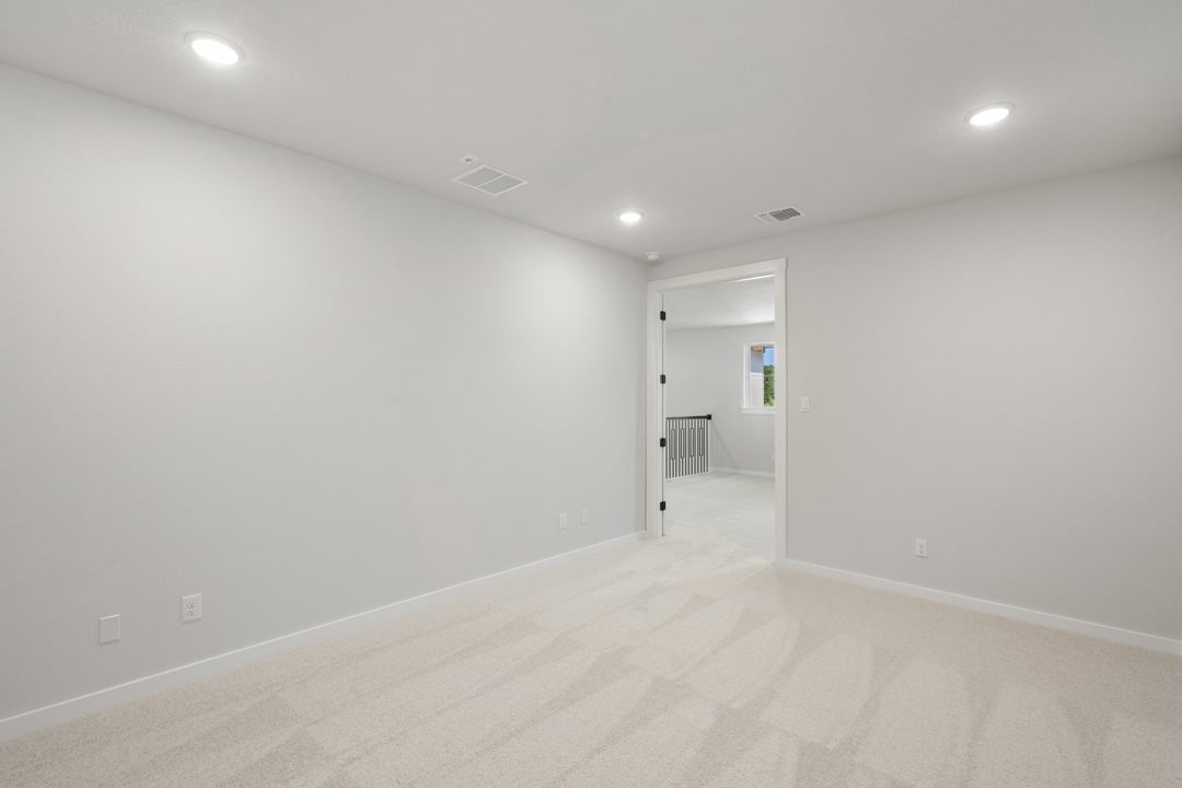 A bright, minimalist room with white walls, a hardwood floor, and recessed lighting, leading to an open doorway.