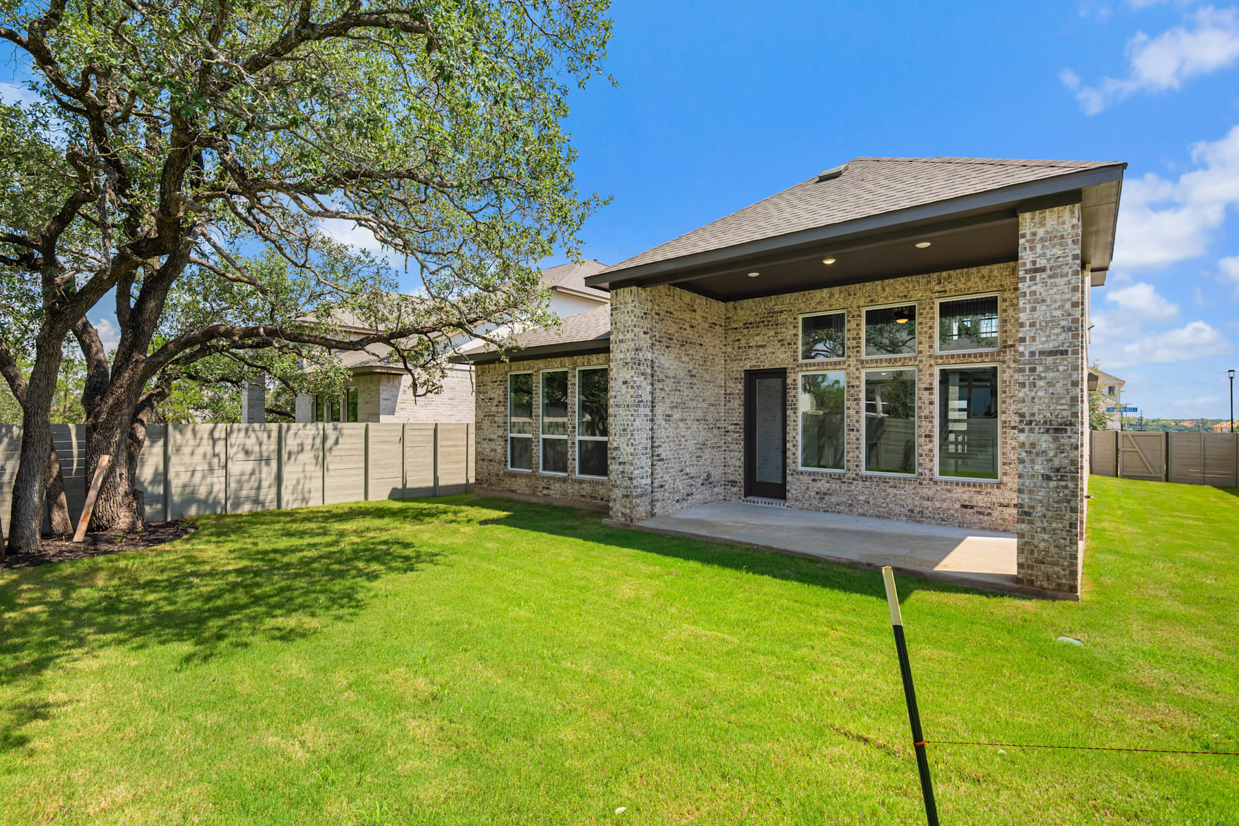 A modern two-story house with a stone exterior, surrounded by a lush green lawn and a large tree in the foreground, set against a clear blue sky.