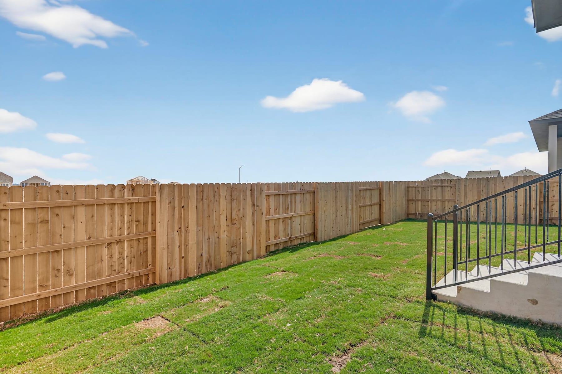 A wooden fence surrounds a grassy backyard with a clear blue sky and fluffy white clouds in the background.