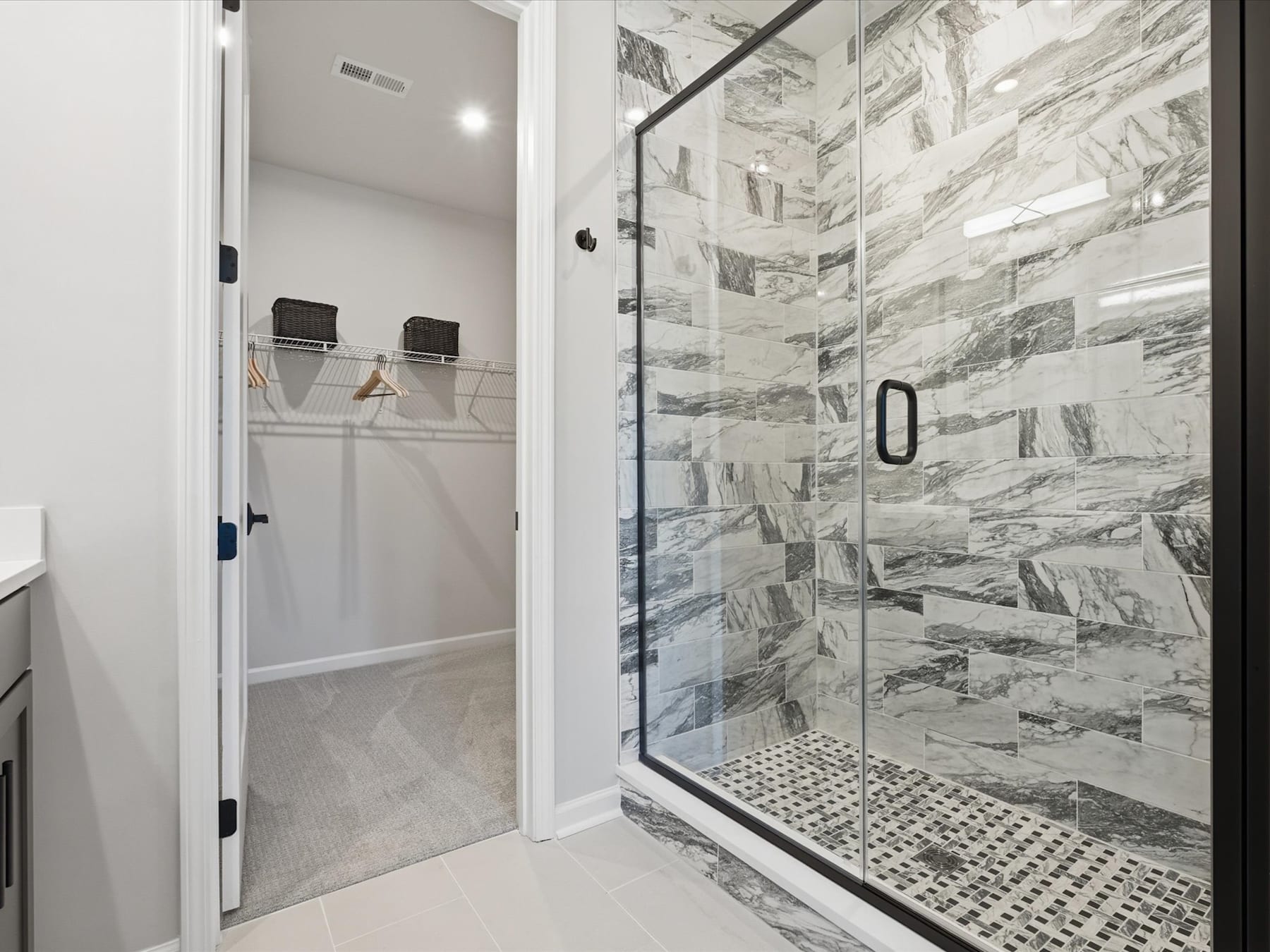 A modern, minimalist bathroom with a glass-enclosed shower, featuring a tiled wall with a striking marble-like pattern and a white door leading to a closet or storage space.