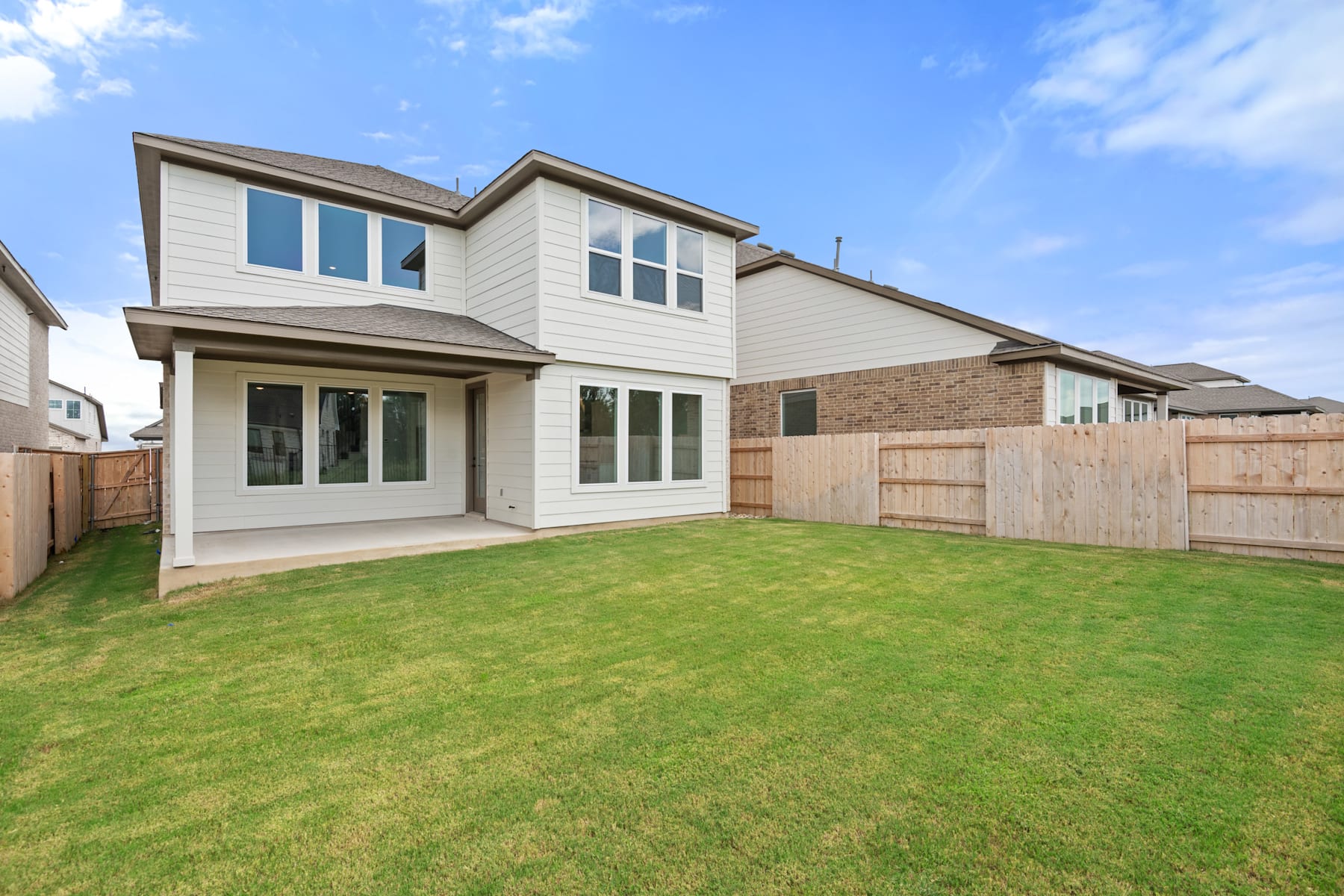 A two-story residential house with a well-manicured lawn and a wooden fence in the foreground, set against a clear blue sky with scattered clouds.