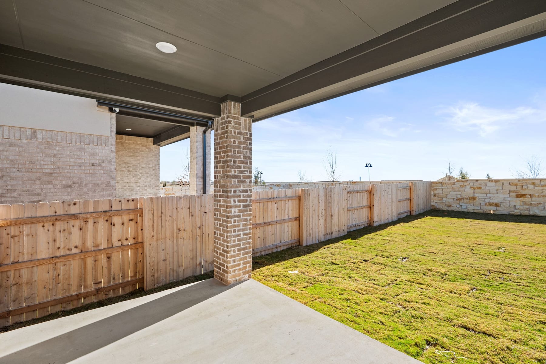 A covered patio with a brick column leads to a grassy backyard surrounded by a wooden fence, with a cloudy sky visible in the background.