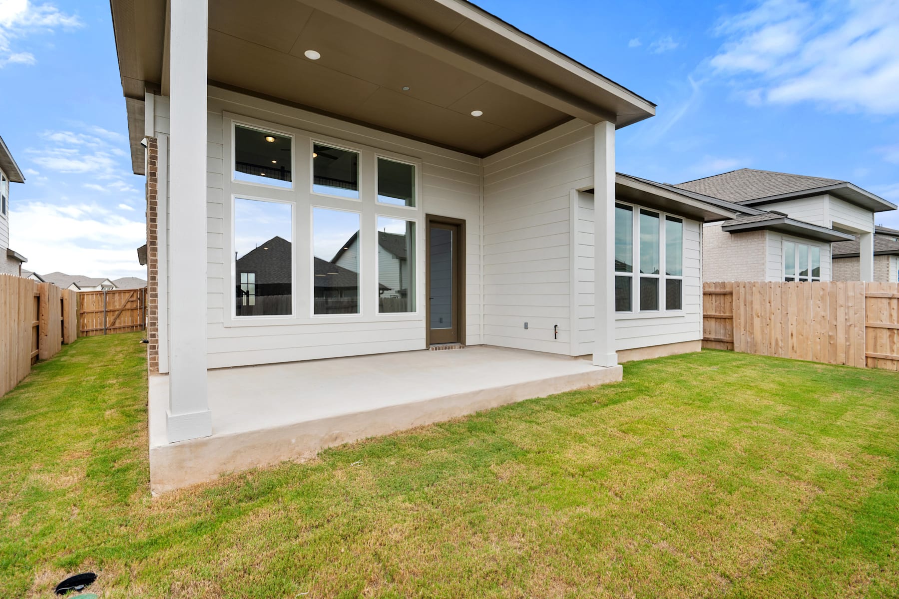 A modern, two-story house with a large grassy yard in the foreground, surrounded by a wooden fence and set against a clear blue sky.