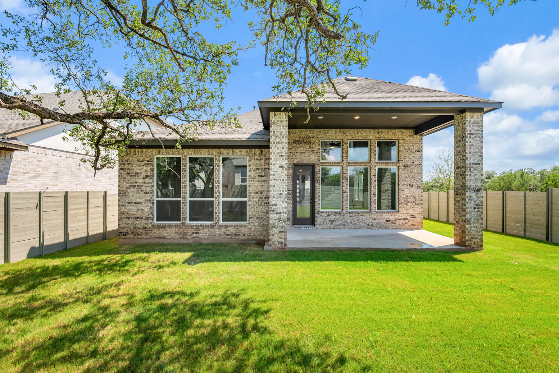 A modern, two-story brick house with large windows and a covered porch sits on a well-manicured lawn surrounded by lush greenery and a clear blue sky.