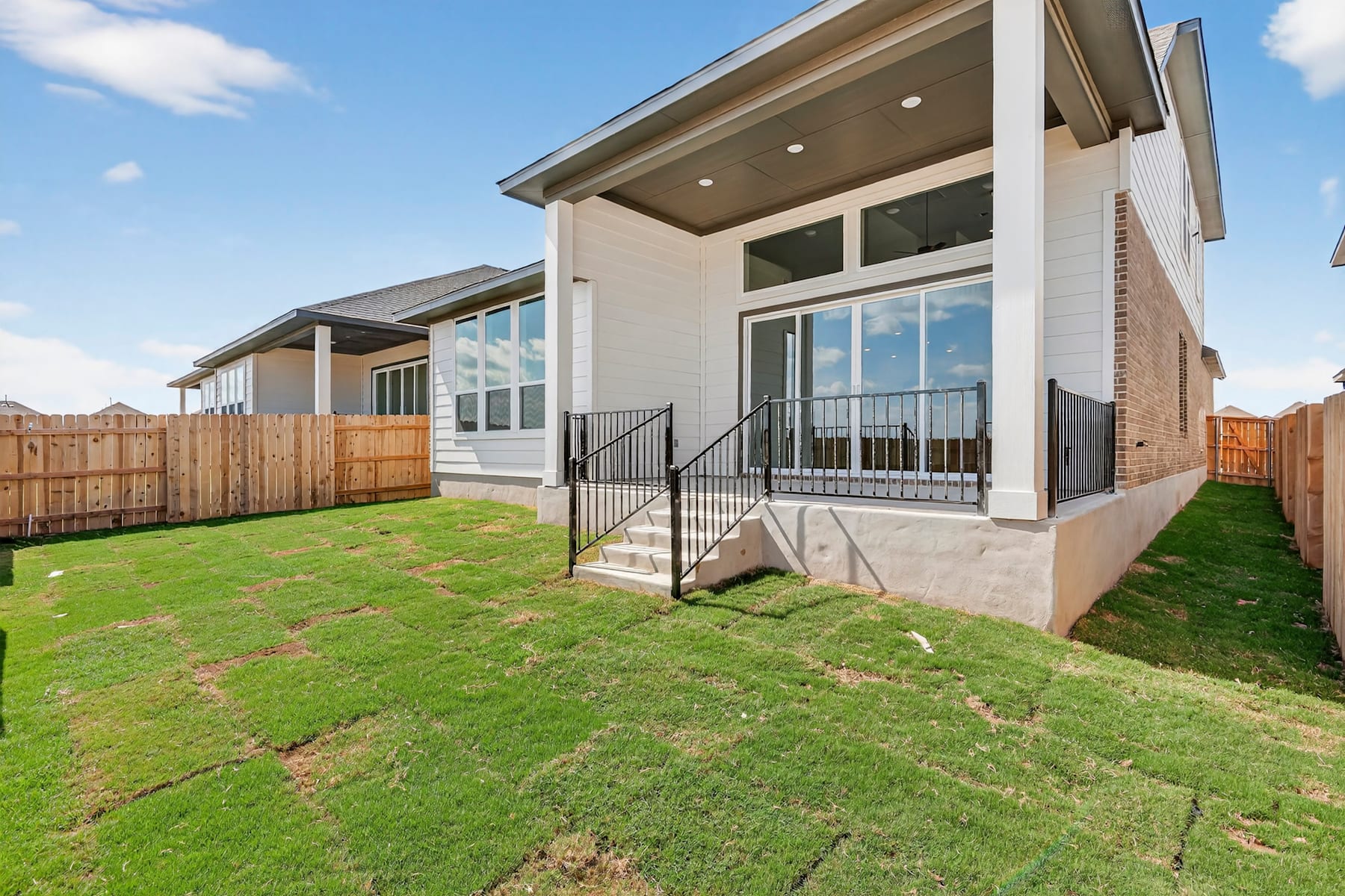 A modern two-story house with a well-manicured lawn and a wooden fence in the foreground, set against a clear blue sky with scattered clouds.
