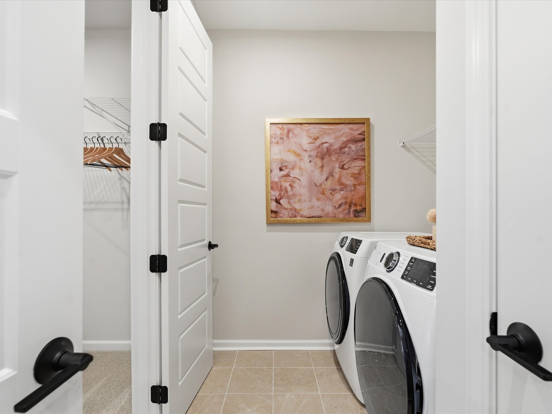 A bright and airy laundry room with a washer and dryer, a framed artwork on the wall, and a closet with white doors.