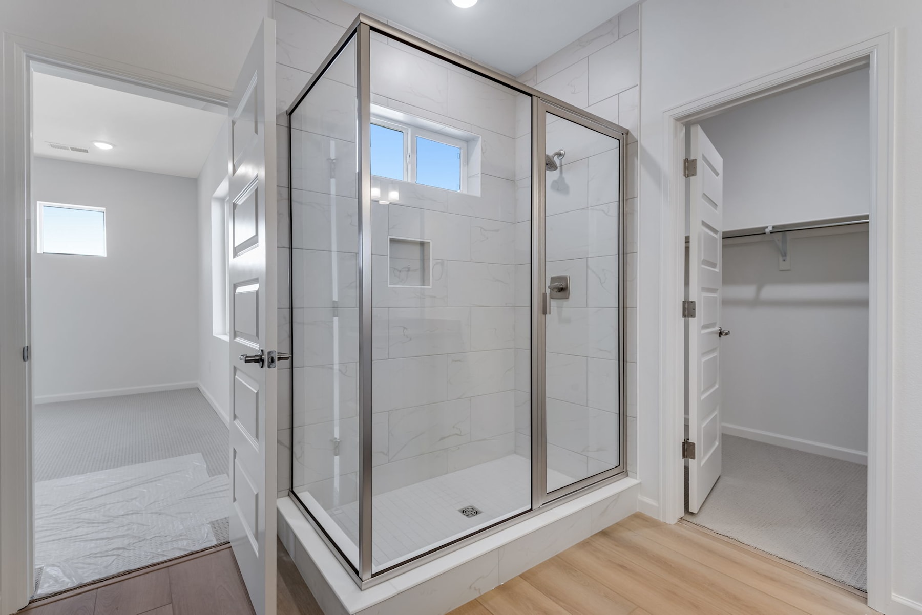 A modern, minimalist bathroom with a large glass-enclosed shower, white tile walls, and a wooden floor.