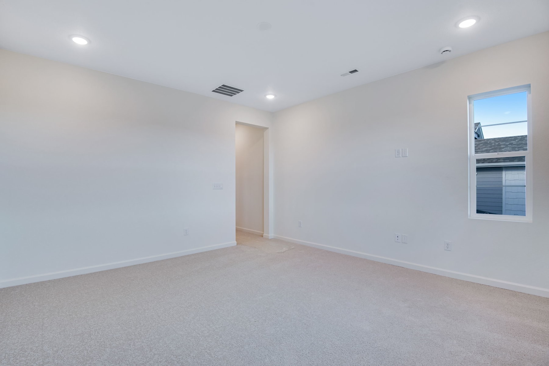 An empty, well-lit room with white walls, a window, and recessed lighting fixtures on the ceiling.