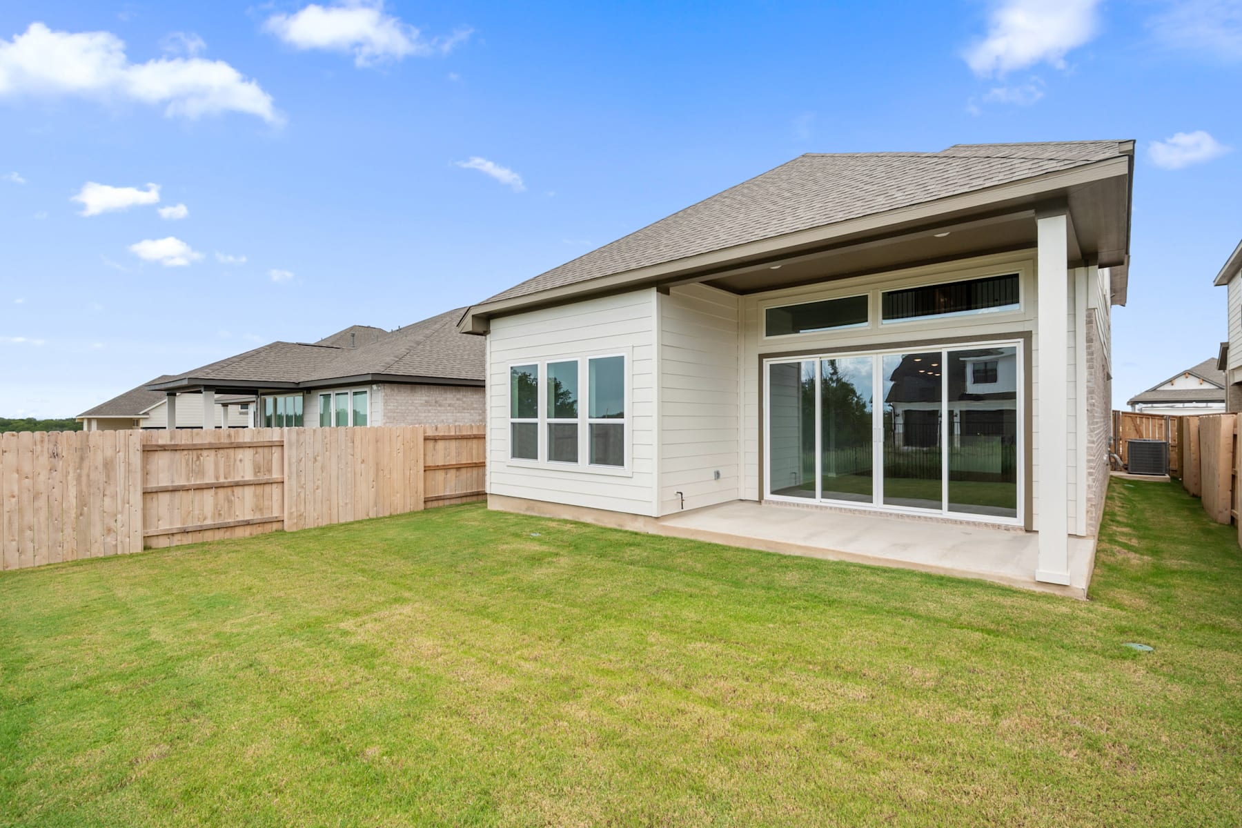 A modern, single-story house with a large grassy yard and a wooden fence in the background, set against a bright blue sky with fluffy white clouds.