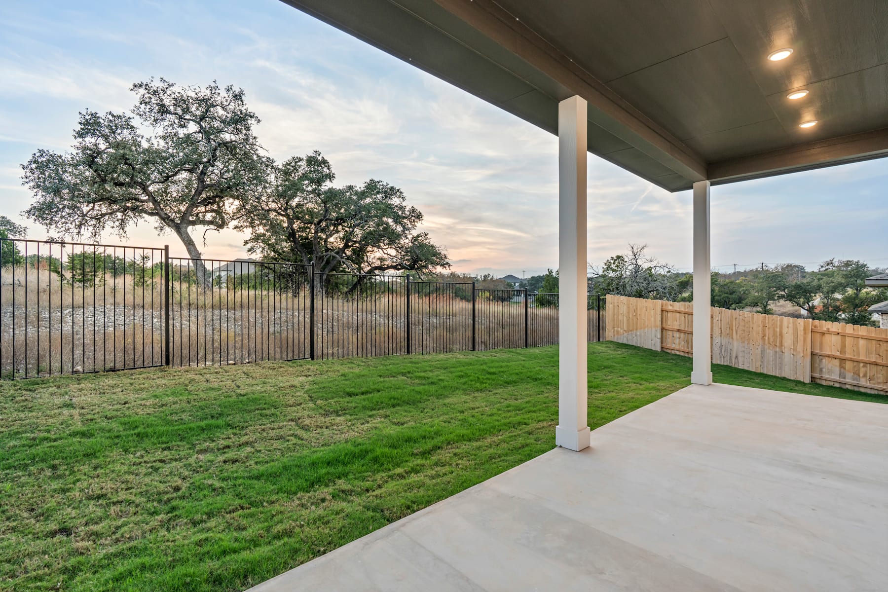 A covered patio with a grassy yard, a wooden fence, and a tree in the background, set against a scenic sky.