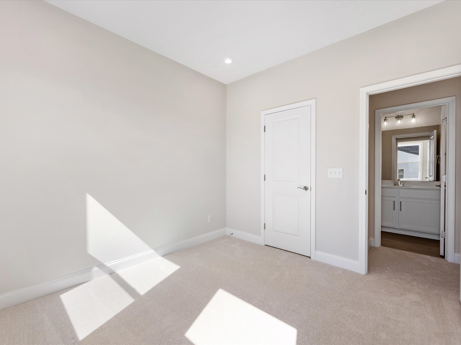 A bright, minimalist bedroom with a white door, neutral-colored walls, and a carpeted floor bathed in natural light from a window.