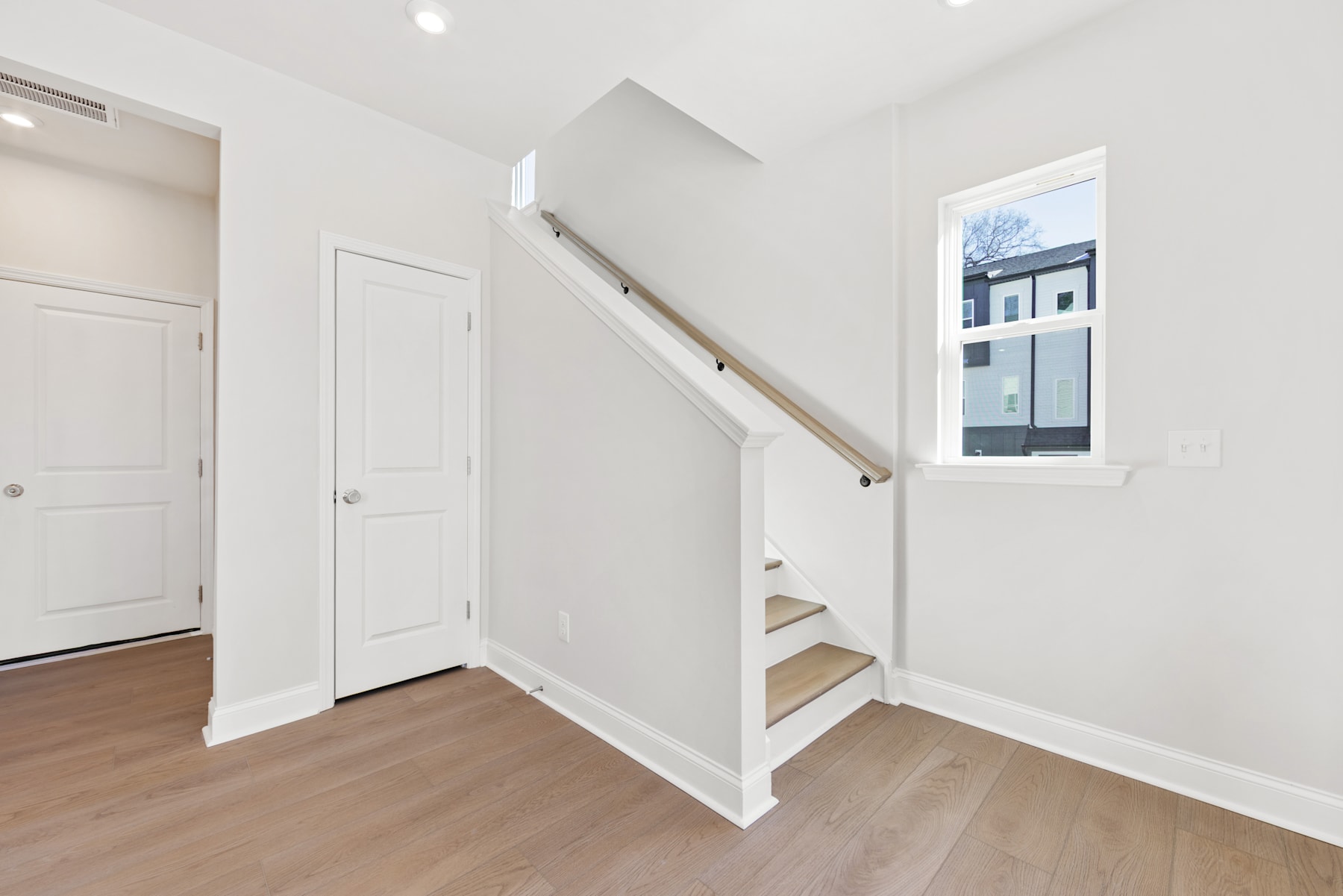 A bright and spacious interior with white walls, wooden floors, and a staircase leading to an upper level. The room features a window, doors, and a minimalist design aesthetic.