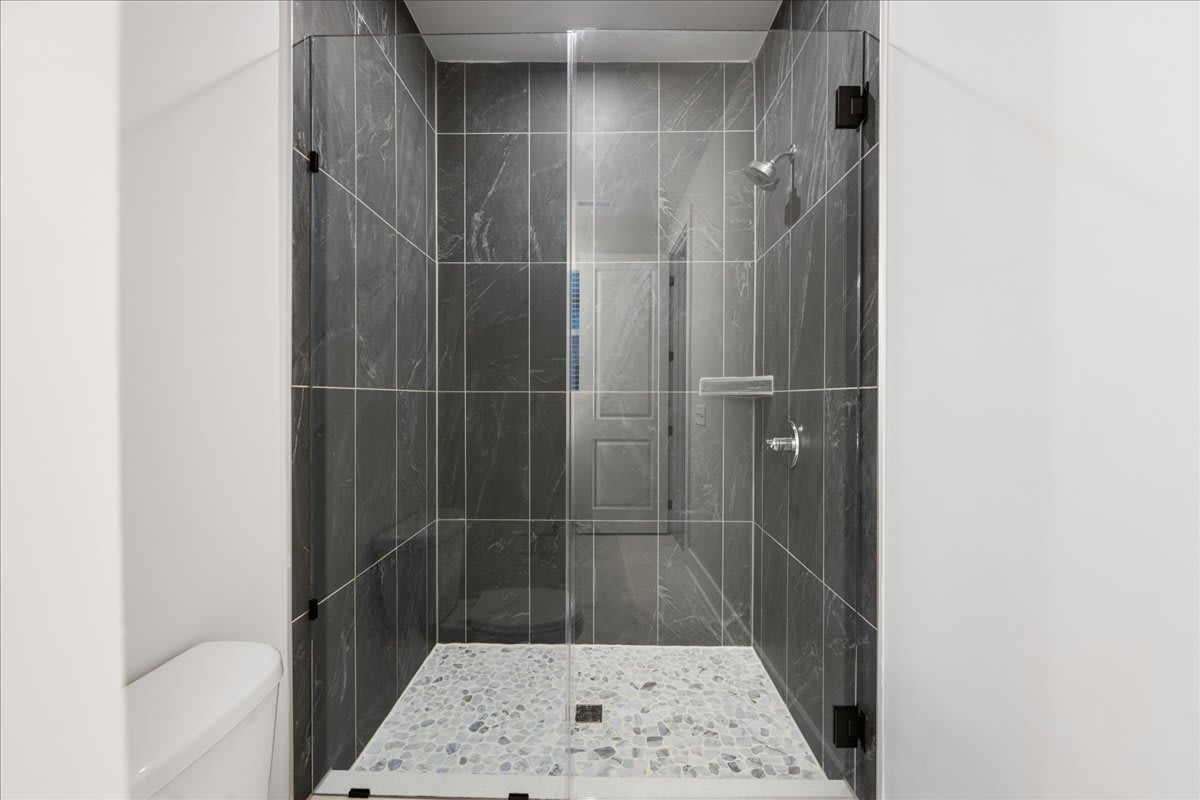 A modern, minimalist bathroom with a glass-enclosed shower stall featuring gray tile walls and a terrazzo-style floor.