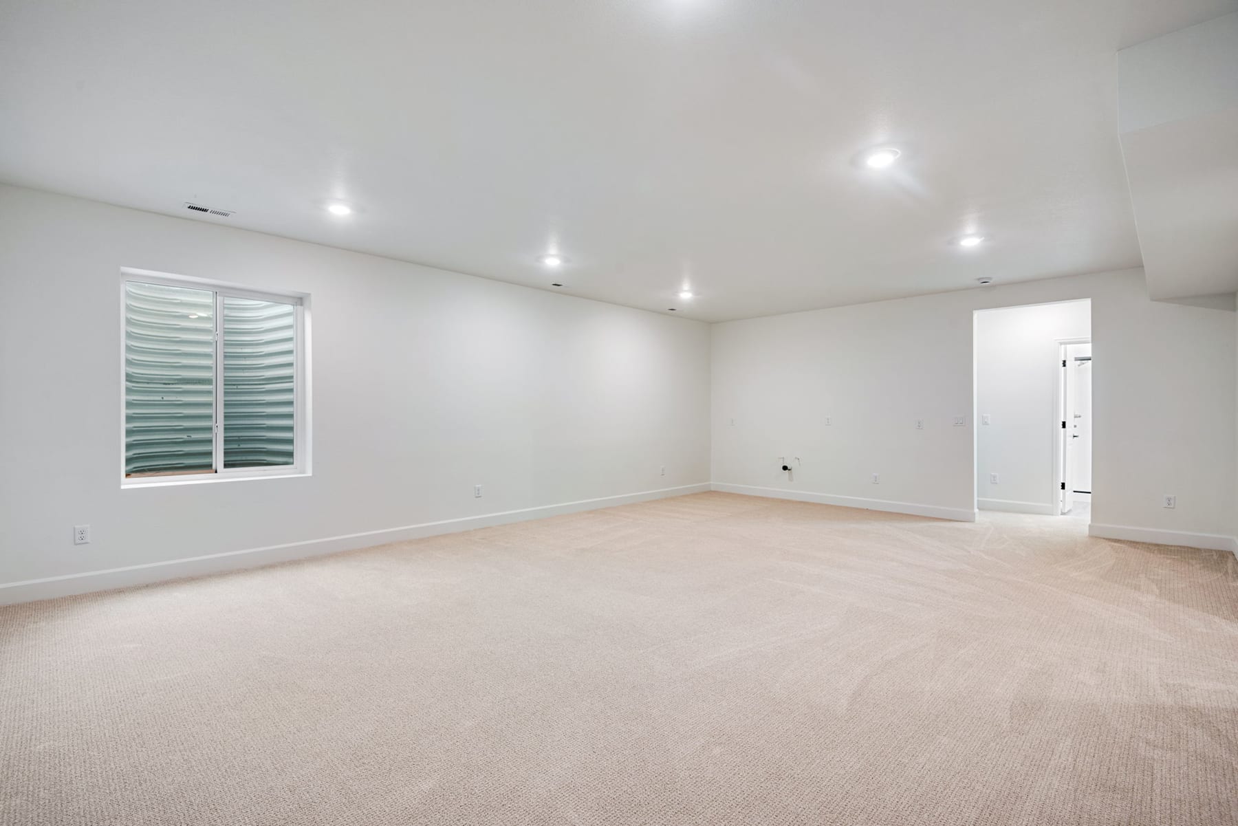 A spacious, well-lit room with a large window, white walls, and a carpeted floor.