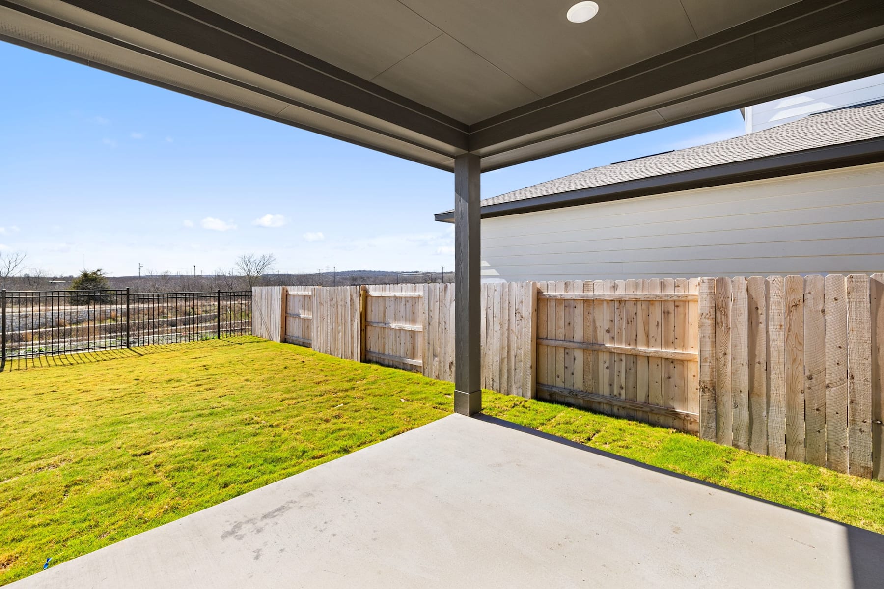 A covered patio with a grassy yard and a wooden fence in the background, under a clear blue sky.