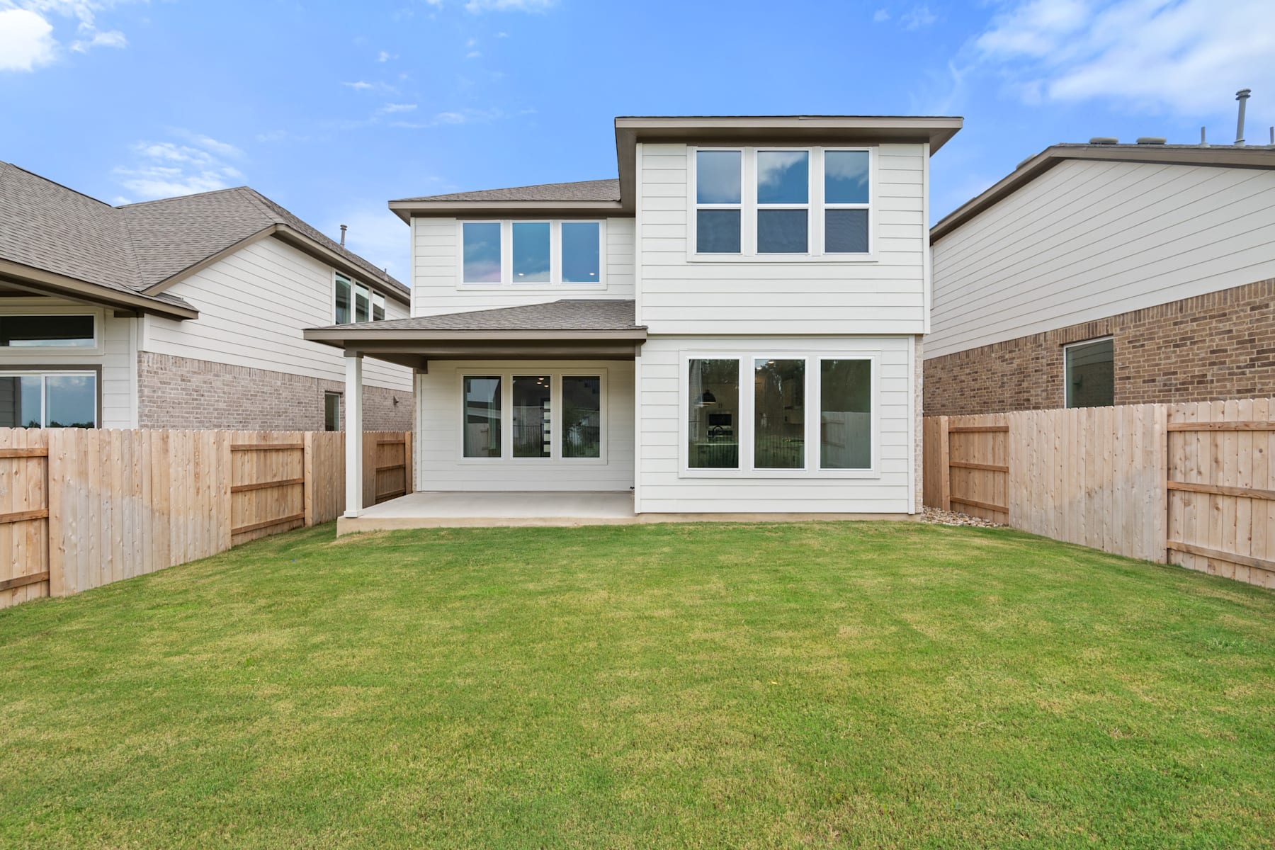 A modern two-story house with a well-manicured lawn in the foreground, surrounded by a wooden fence and other residential buildings in the background.