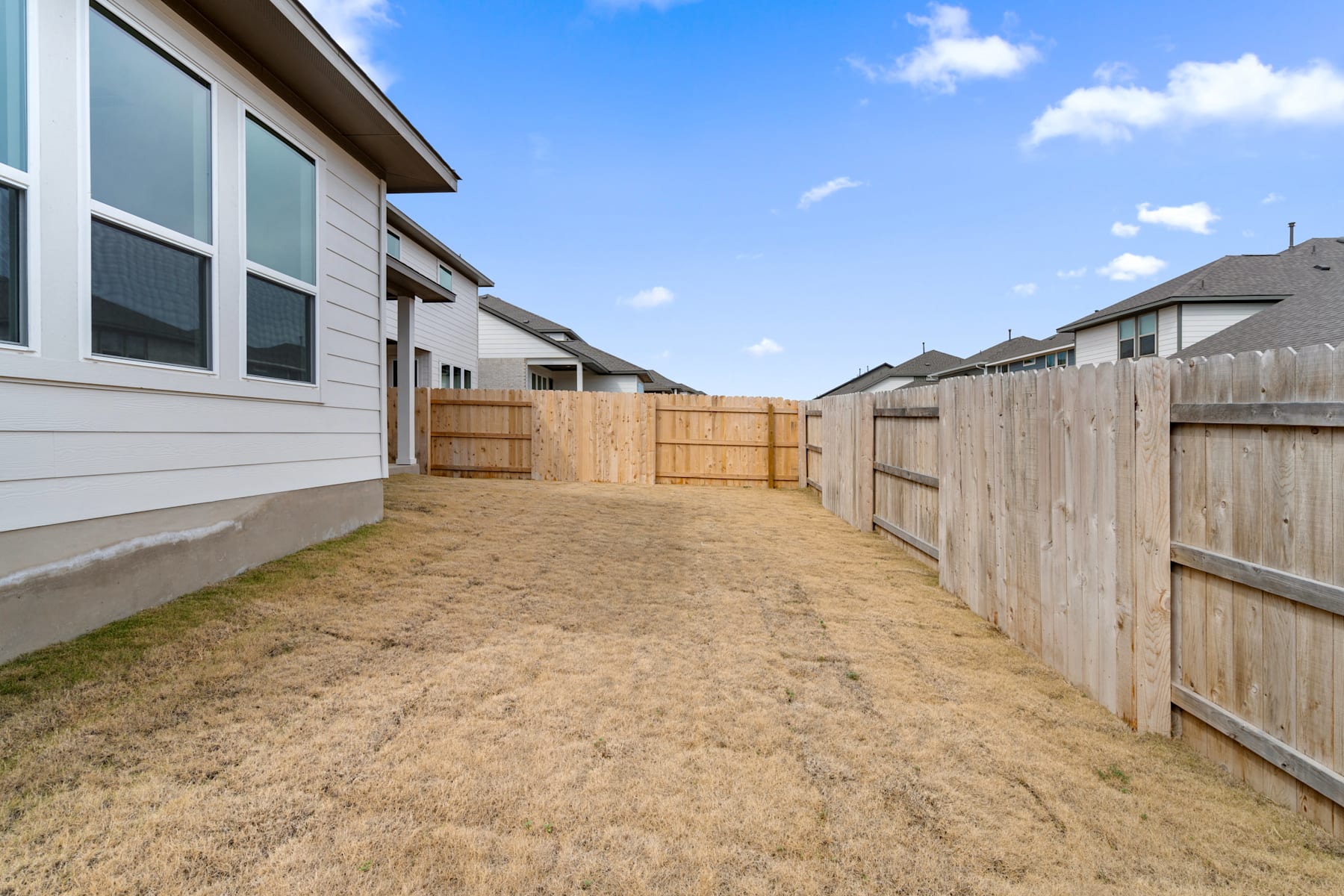 A dirt path leads between wooden fences, with a residential building visible in the background under a blue sky with scattered clouds.
