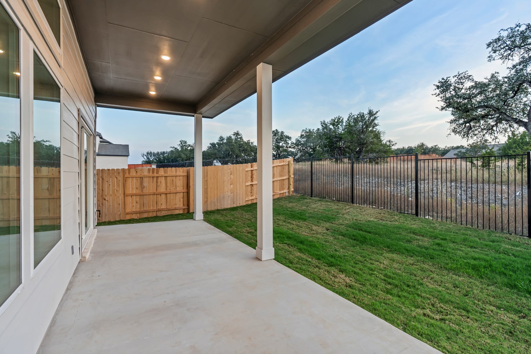 A covered patio with a concrete floor leads to a grassy backyard surrounded by a wooden fence, with trees and a clear sky visible in the background.