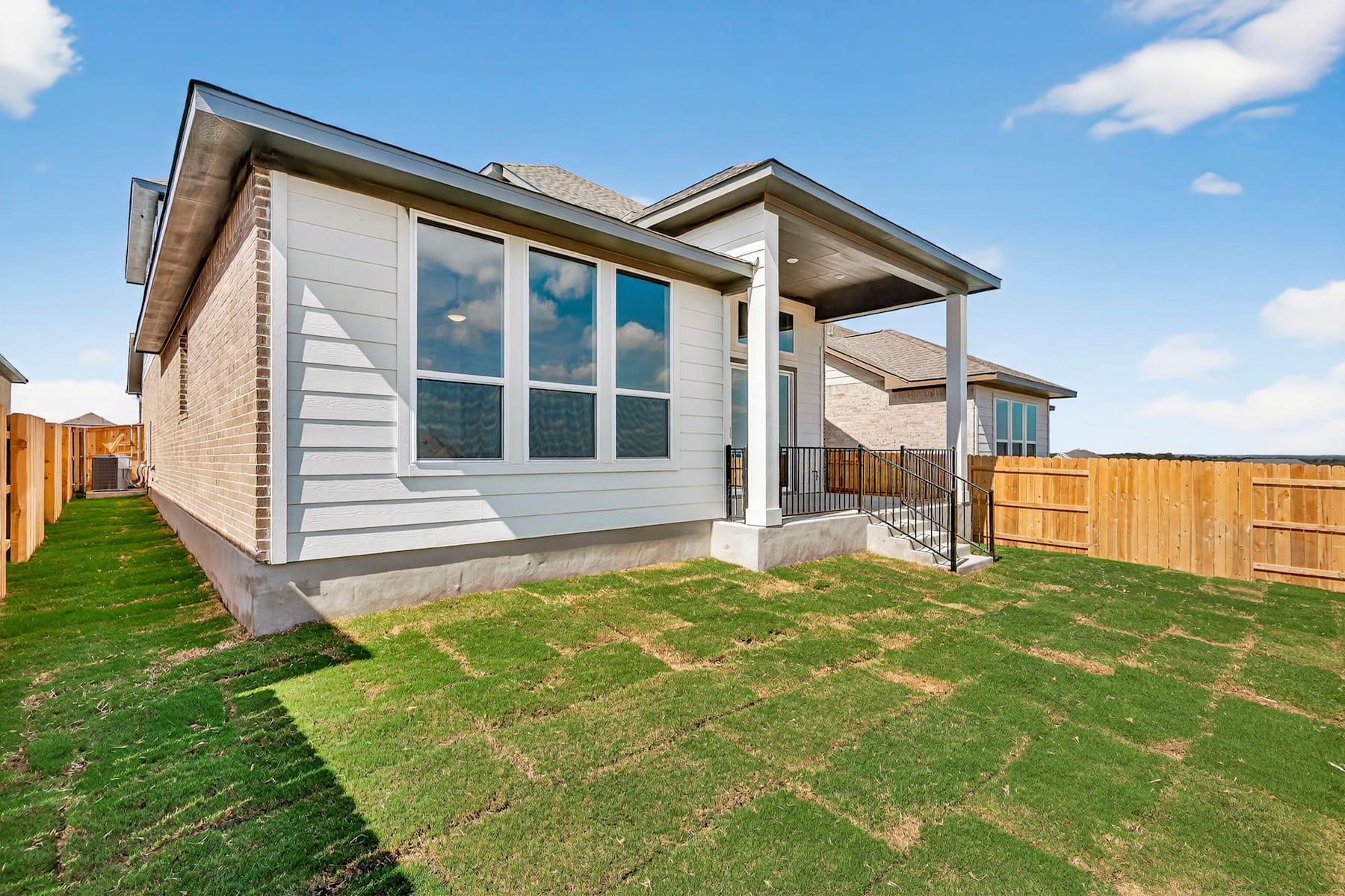 A modern two-story house with a well-manicured lawn and a wooden fence in the foreground, set against a clear blue sky with scattered clouds.