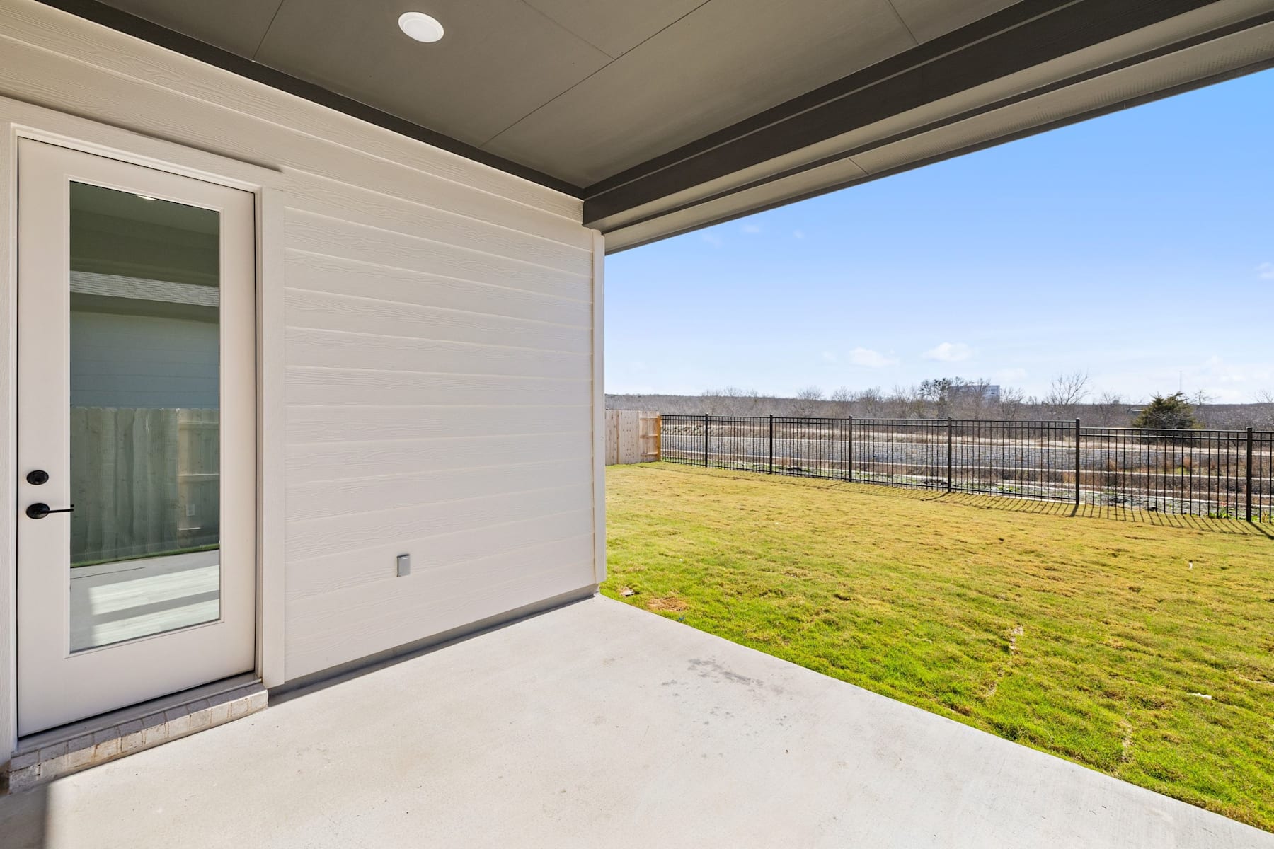 A modern, white-walled patio with a glass door leading to a grassy, fenced-in backyard under a clear blue sky.