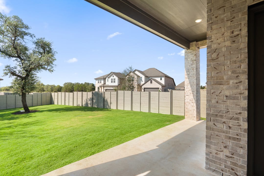 A well-manicured lawn surrounded by a brick wall and fencing, with a two-story house visible in the background against a clear blue sky.