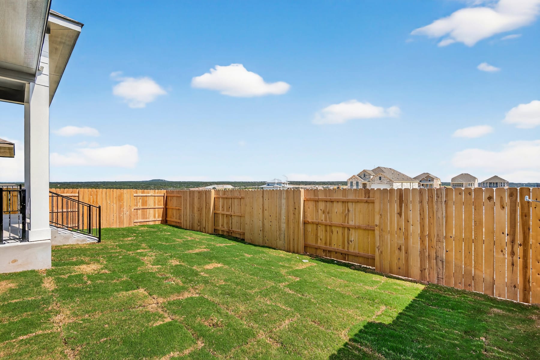 A wooden fence surrounds a grassy backyard, with houses visible in the background against a blue sky with fluffy white clouds.