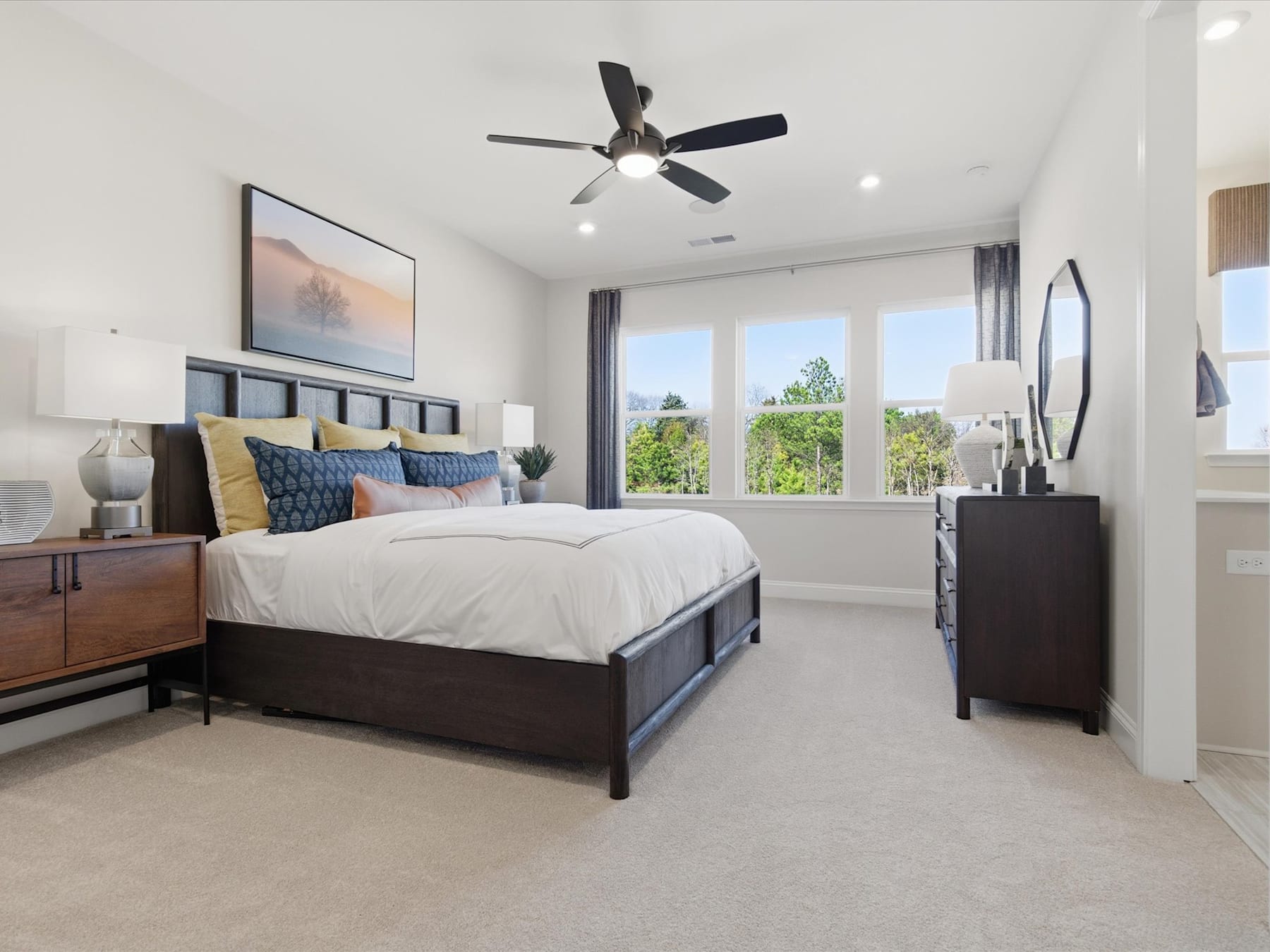 A spacious and well-furnished bedroom with a large bed, dark wood furniture, and a ceiling fan, set against a backdrop of large windows overlooking a scenic outdoor view.